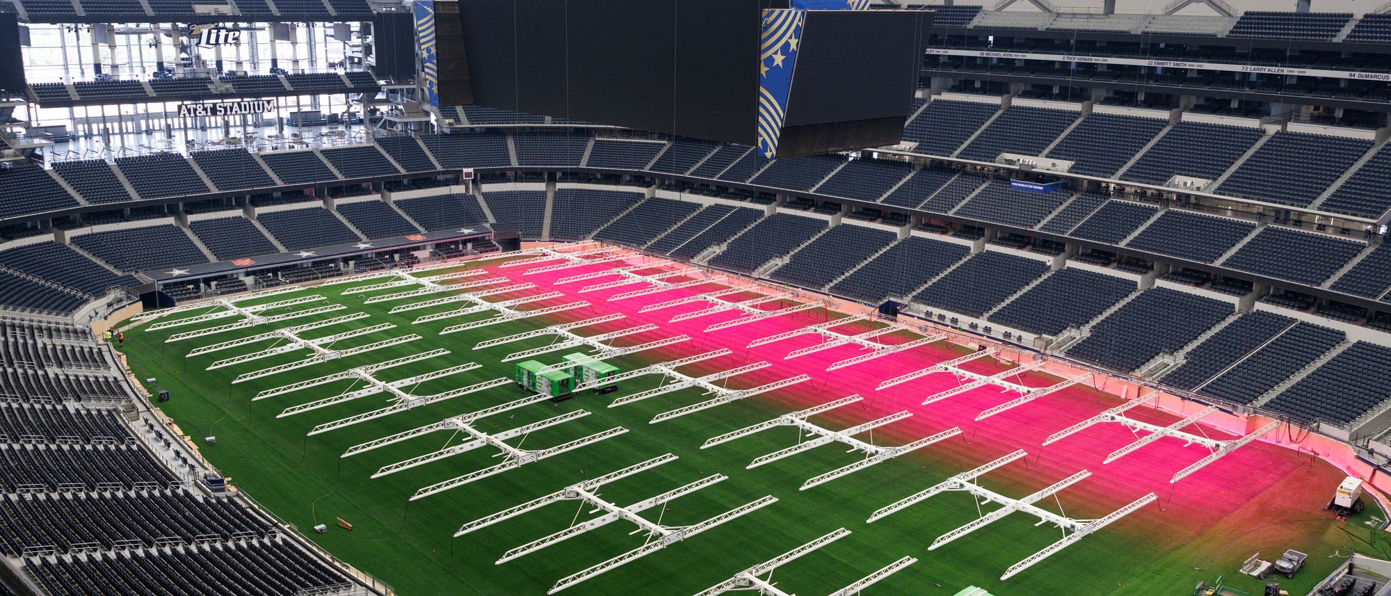 Ein gro&szlig;es, leeres Stadion mit einem gr&uuml;nen Spielfeld, das teilweise von pinkfarbenem Licht beleuchtet wird. &Uuml;ber dem Spielfeld sind wei&szlig;e Metallstrukturen verteilt. Im Hintergrund sind gro&szlig;e Bildschirme und Werbetafeln sichtbar. Das Stadion ist von leeren Sitzreihen umgeben.