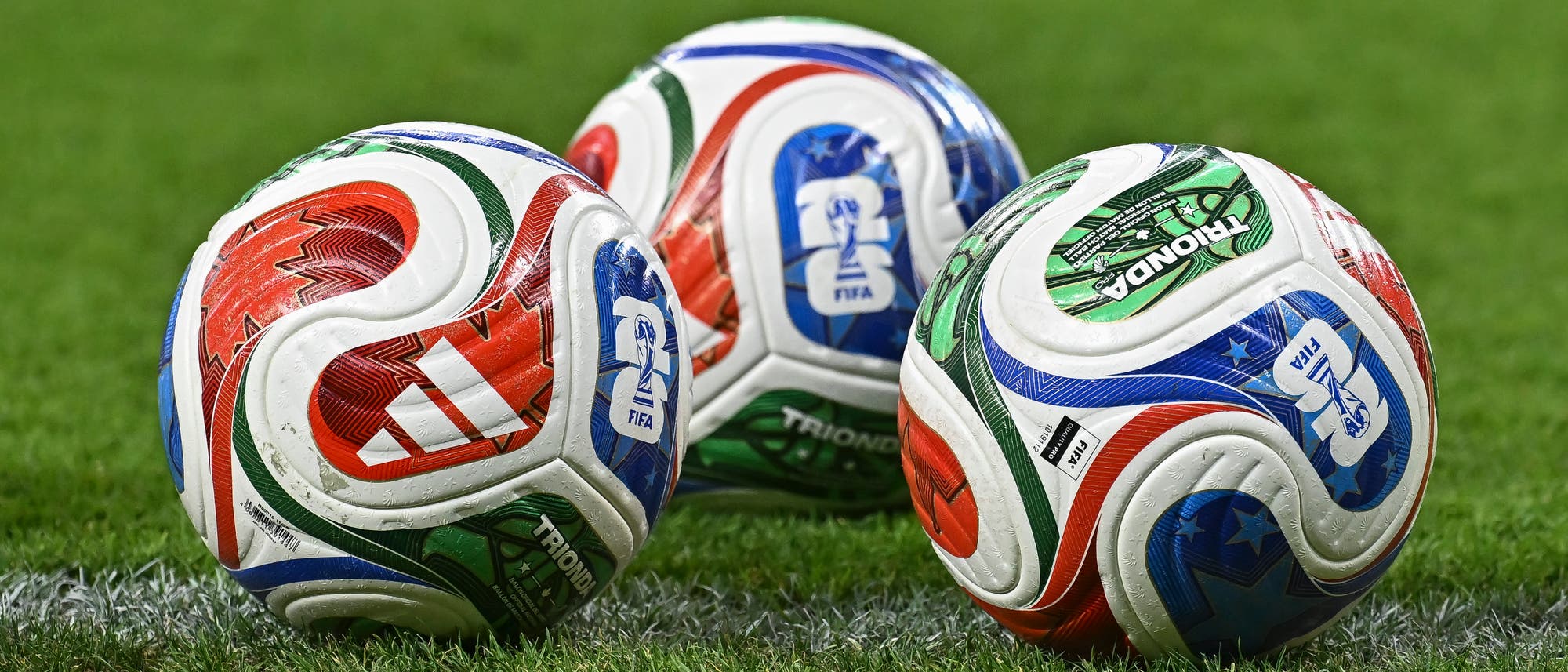 Drei bunte Fu&szlig;b&auml;lle mit FIFA-Logo und Adidas-Branding liegen auf einem gr&uuml;nen Fu&szlig;ballfeld. Die B&auml;lle zeigen ein Design in Rot, Blau und Gr&uuml;n.