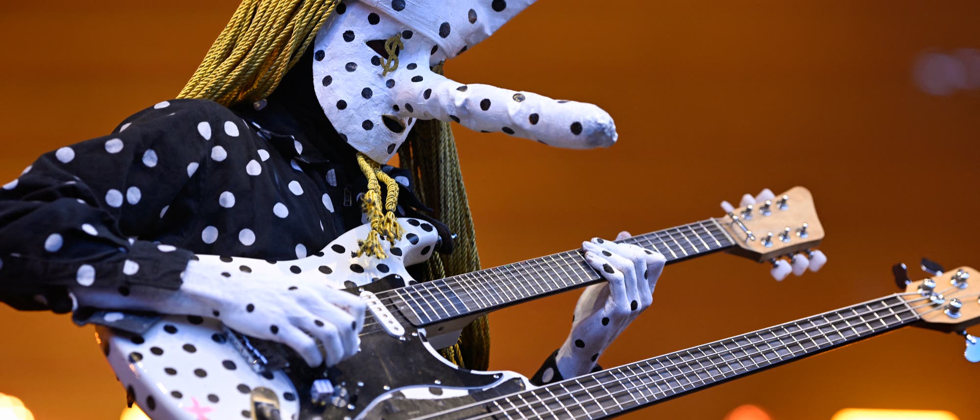 Eine Person in einem auffälligen Kostüm mit großen, gepunkteten Elementen spielt eine doppelte E-Gitarre. Das Kostüm ist schwarz-weiß gepunktet, und die Gitarre hat ein ähnliches Muster. Die Szene ist in warmes Licht getaucht. Eine Person in einem auffälligen Kostüm mit großen, gepunkteten Elementen spielt eine doppelte E-Gitarre. Das Kostüm ist schwarz-weiß gepunktet, und die Gitarre hat ein ähnliches Muster. Die Szene ist in warmes Licht getaucht.