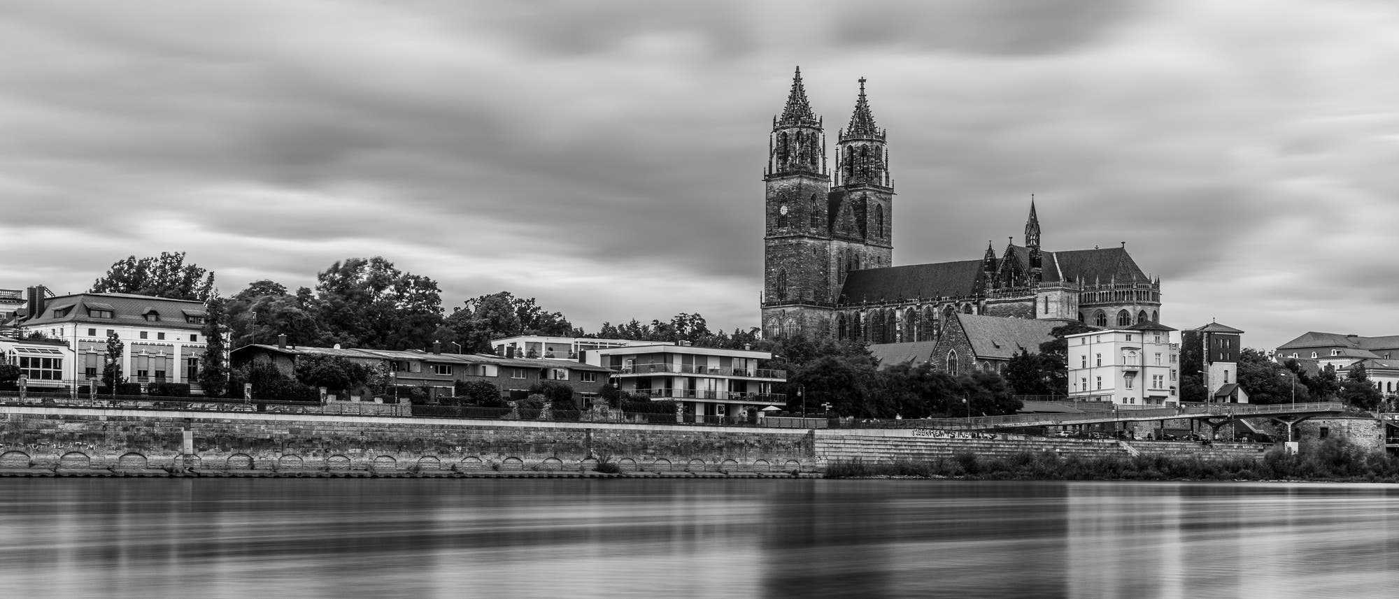 Schwarz-Weiß-Fotografie einer historischen Kathedrale mit zwei hohen Türmen, die sich majestätisch über eine Reihe moderner und traditioneller Gebäude am Flussufer erhebt. Der Himmel ist bewölkt, und das Wasser im Vordergrund spiegelt die ruhige Atmosphäre wider. Schwarz-Weiß-Fotografie einer historischen Kathedrale mit zwei hohen Türmen, die sich majestätisch über eine Reihe moderner und traditioneller Gebäude am Flussufer erhebt. Der Himmel ist bewölkt, und das Wasser im Vordergrund spiegelt die ruhige Atmosphäre wider.