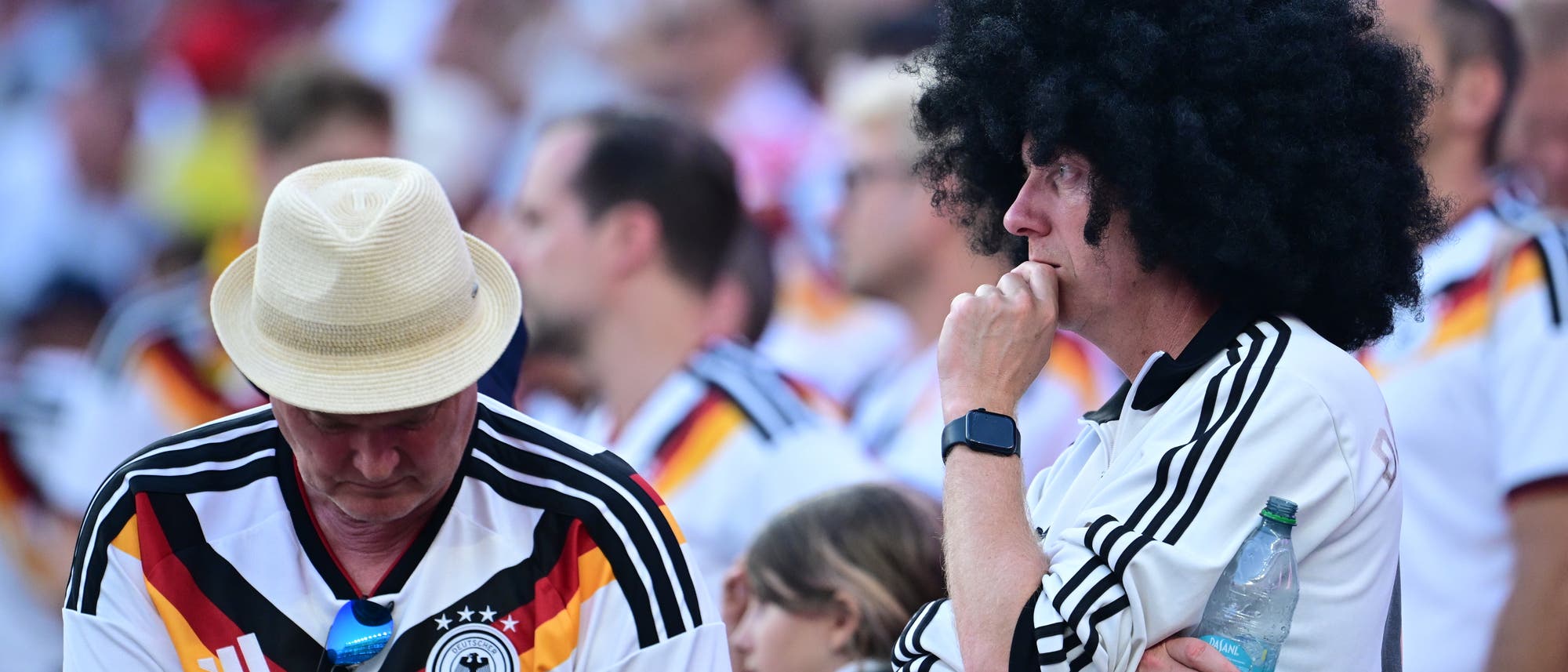 Zwei Personen in Fu&szlig;balltrikots der deutschen Nationalmannschaft stehen in einem Stadion. Eine Person tr&auml;gt einen Strohhut und schaut nach unten, w&auml;hrend die andere eine gro&szlig;e, schwarze Per&uuml;cke tr&auml;gt und nachdenklich wirkt. Im Hintergrund sind weitere Zuschauer zu sehen.
