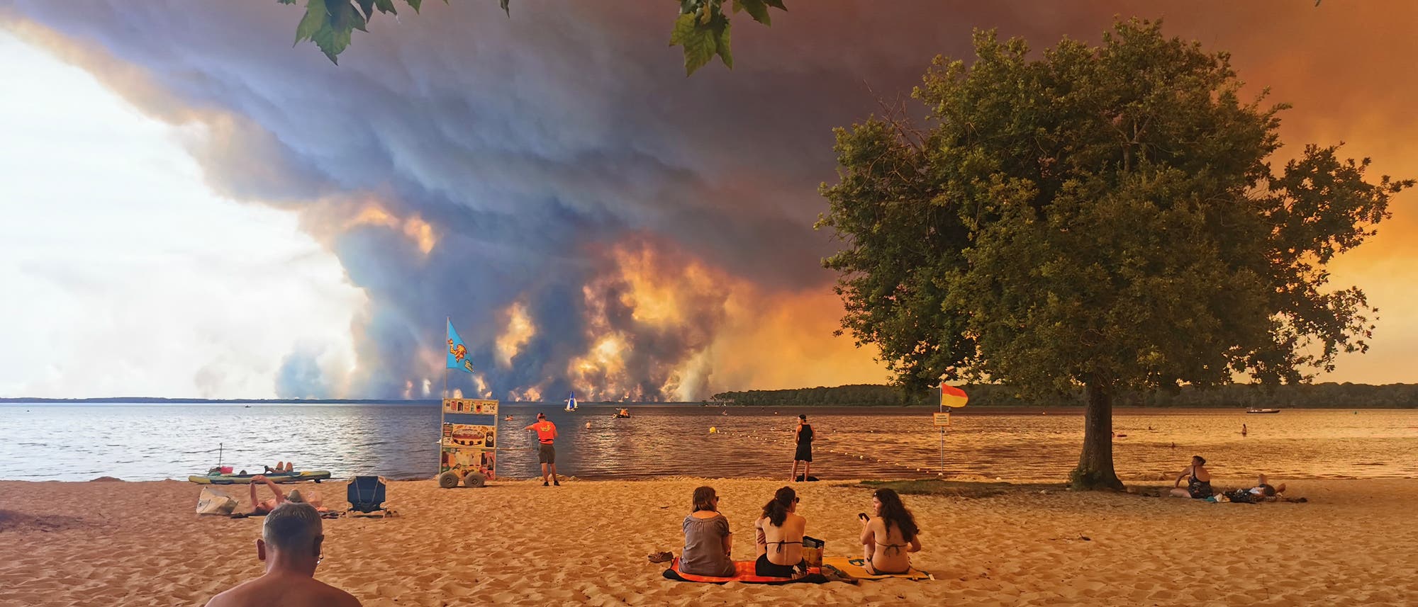 Strand mit mehreren Menschen, die auf Handtüchern sitzen und in Richtung eines großen Waldbrands am Horizont blicken. Der Himmel ist von dichten Rauchwolken bedeckt, die sich über das Wasser erstrecken. Strand mit mehreren Menschen, die auf Handtüchern sitzen und in Richtung eines großen Waldbrands am Horizont blicken. Der Himmel ist von dichten Rauchwolken bedeckt, die sich über das Wasser erstrecken.