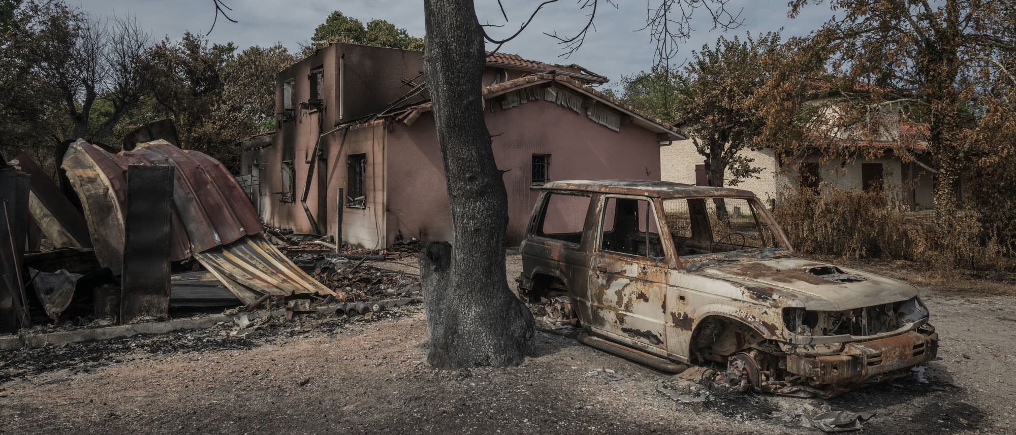 Ein ausgebranntes Auto steht neben einem verkohlten Baum vor einem stark beschädigten Haus mit verbranntem Dach. Die Umgebung zeigt Anzeichen eines kürzlichen Feuers, mit verbrannten Bäumen und Trümmern. Der Himmel ist bewölkt, was eine düstere Atmosphäre schafft. Ein ausgebranntes Auto steht neben einem verkohlten Baum vor einem stark beschädigten Haus mit verbranntem Dach. Die Umgebung zeigt Anzeichen eines kürzlichen Feuers, mit verbrannten Bäumen und Trümmern. Der Himmel ist bewölkt, was eine düstere Atmosphäre schafft.