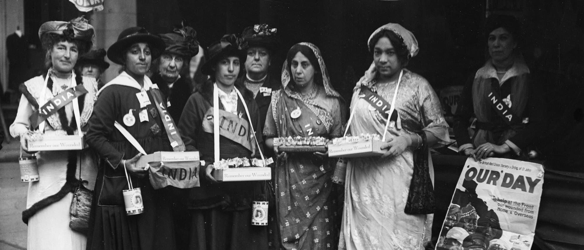 Eine Gruppe von Frauen steht nebeneinander und h&auml;lt Schachteln mit der Aufschrift &bdquo;Remember our wounded&ldquo;. Sie tragen Sch&auml;rpen mit der Aufschrift &bdquo;India&ldquo; und sind in verschiedenen traditionellen und westlichen Kleidungsstilen gekleidet. Im Hintergrund sind Plakate mit der Aufschrift &bdquo;Our Day&ldquo; zu sehen, die auf eine Veranstaltung am Donnerstag, den 19. Oktober, hinweisen. Die Szene scheint eine Wohlt&auml;tigkeitsveranstaltung oder eine Spendenaktion zu sein.