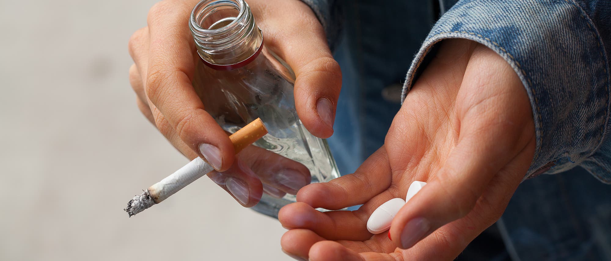 Eine Person in einer Jeansjacke hält in der einen Hand eine brennende Zigarette und eine leere Glasflasche, in der anderen Hand zwei weiße Tabletten. Die Szene vermittelt den Eindruck von Substanzmissbrauch. Eine Person in einer Jeansjacke hält in der einen Hand eine brennende Zigarette und eine leere Glasflasche, in der anderen Hand zwei weiße Tabletten. Die Szene vermittelt den Eindruck von Substanzmissbrauch.