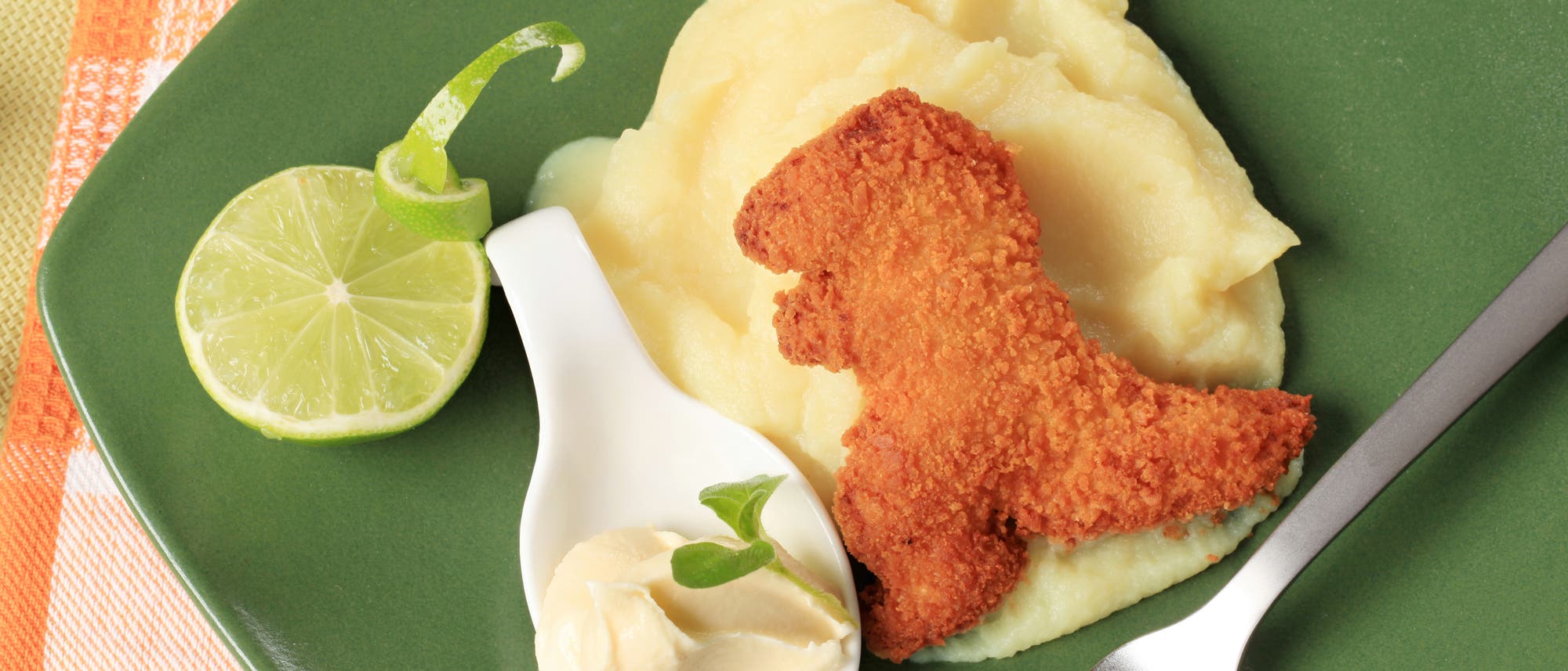 Ein grüner Teller mit Kartoffelpüree, darauf ein paniertes Stück Fleisch in Form eines Dinosauriers. Daneben eine weiße Löffelschale mit einem Klecks Butter und einem kleinen grünen Blatt. Eine halbe Limettenscheibe liegt am Rand. Eine Gabel ist am rechten Rand des Tellers platziert. Der Hintergrund zeigt ein orange-weiß kariertes Tischtuch. Ein grüner Teller mit Kartoffelpüree, darauf ein paniertes Stück Fleisch in Form eines Dinosauriers. Daneben eine weiße Löffelschale mit einem Klecks Butter und einem kleinen grünen Blatt. Eine halbe Limettenscheibe liegt am Rand. Eine Gabel ist am rechten Rand des Tellers platziert. Der Hintergrund zeigt ein orange-weiß kariertes Tischtuch.