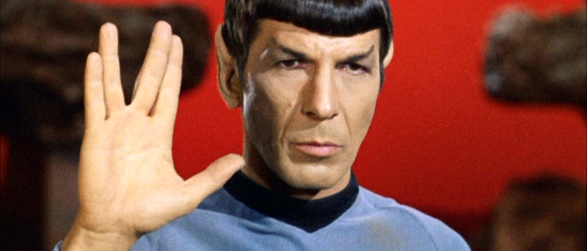 Ein Mann in einem blauen Uniformhemd zeigt den vulkanischen Gruß mit gespreizten Fingern vor einem roten Hintergrund. Die Person hat spitze Ohren, schwarze Haare und einen ernsten Gesichtsausdruck.