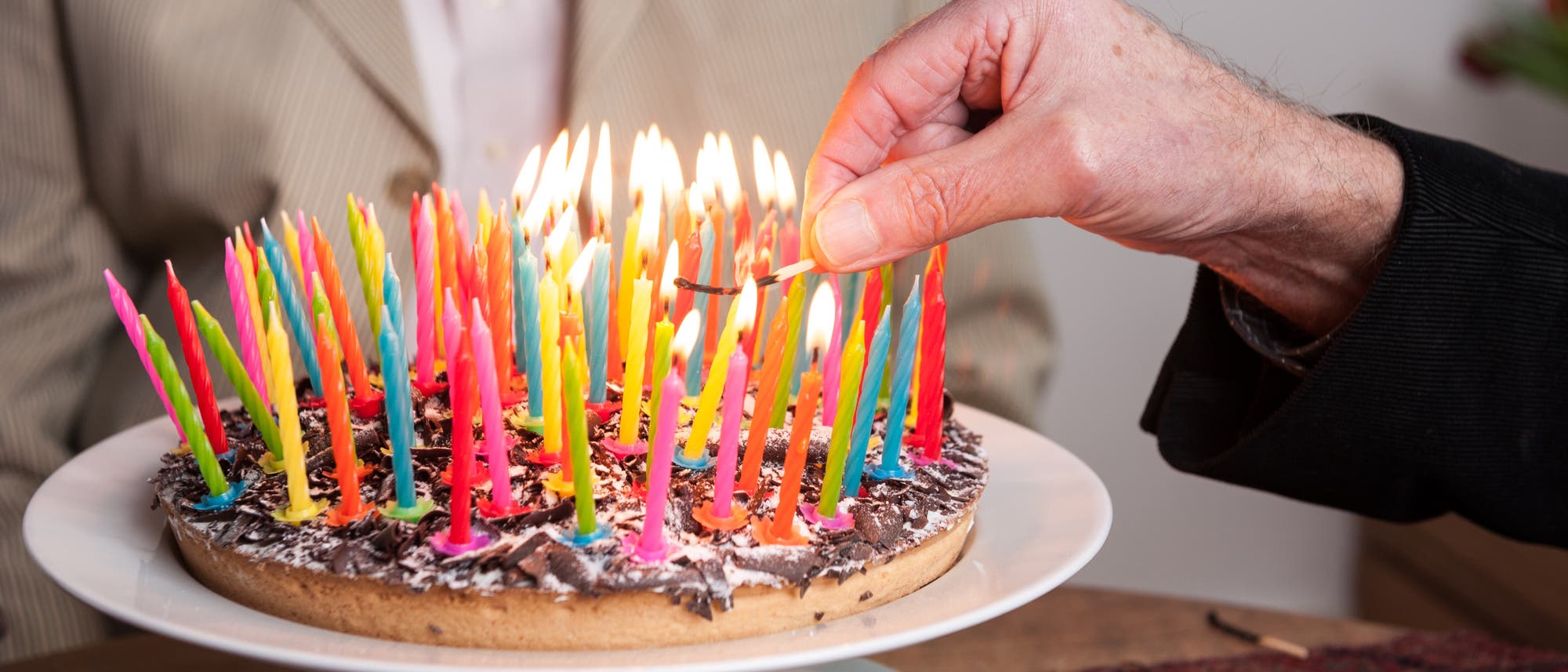 Eine Hand zündet bunte Kerzen auf einem Geburtstagskuchen an. Der Kuchen ist mit Schokoladenraspeln dekoriert und steht auf einem weißen Teller. Im Hintergrund ist eine Person in einem gestreiften Anzug zu sehen. Die Szene vermittelt eine festliche Atmosphäre.