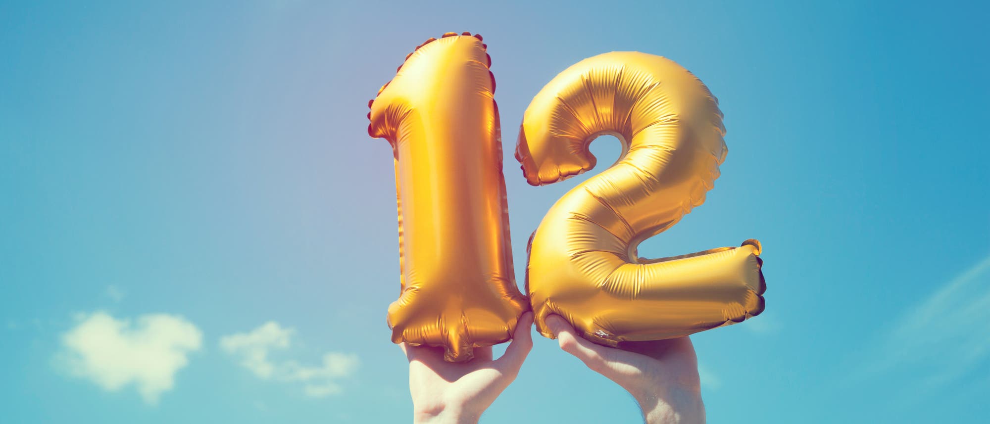 Zwei H&auml;nde halten goldene Ballons in Form der Zahl 12 vor einem klaren blauen Himmel mit vereinzelten Wolken. Die Szene vermittelt ein Gef&uuml;hl von Feierlichkeit und Freude.