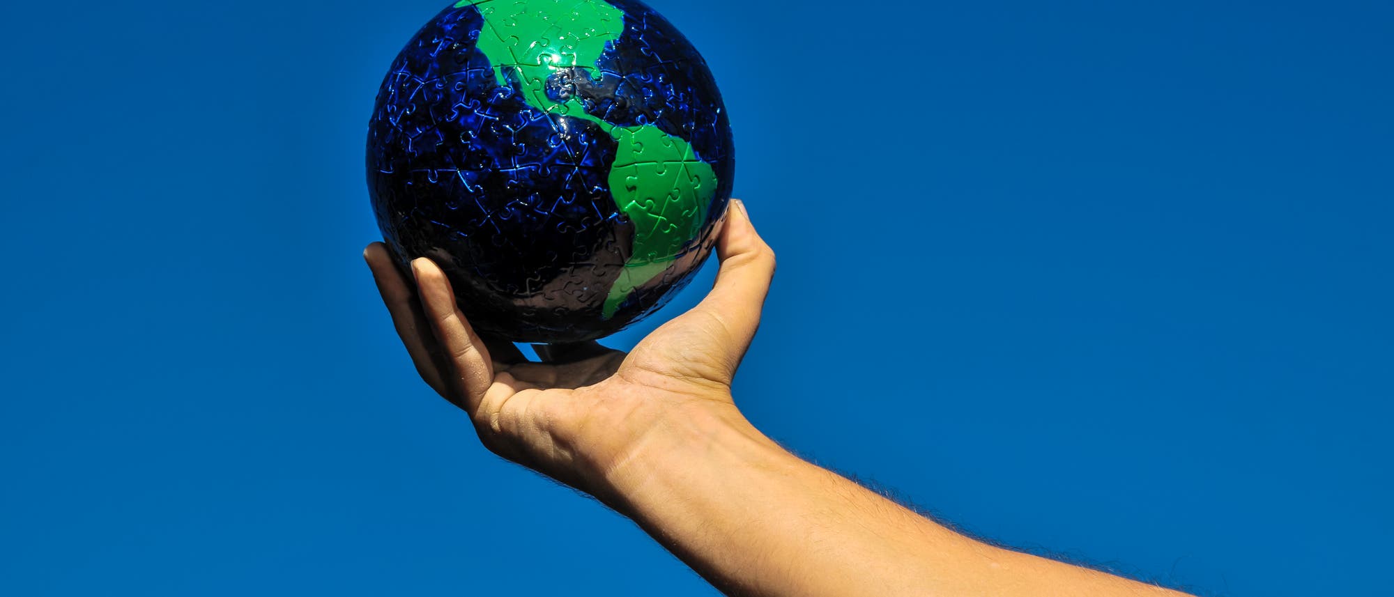 Eine Hand hält einen Puzzleball, der die Erde darstellt, vor einem klaren blauen Himmel. Der Ball zeigt Kontinente in Grün und Ozeane in Blau. Die Szene symbolisiert den Schutz und die Verantwortung für unseren Planeten. Eine Hand hält einen Puzzleball, der die Erde darstellt, vor einem klaren blauen Himmel. Der Ball zeigt Kontinente in Grün und Ozeane in Blau. Die Szene symbolisiert den Schutz und die Verantwortung für unseren Planeten.