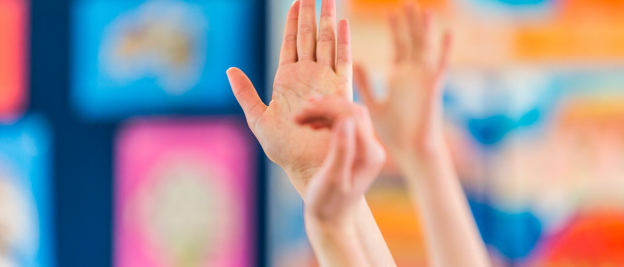 Mehrere Hände sind in einem Klassenzimmer in die Luft gestreckt, als ob sie sich melden. Der Hintergrund ist unscharf und zeigt bunte Poster oder Tafeln. Mehrere Hände sind in einem Klassenzimmer in die Luft gestreckt, als ob sie sich melden. Der Hintergrund ist unscharf und zeigt bunte Poster oder Tafeln.