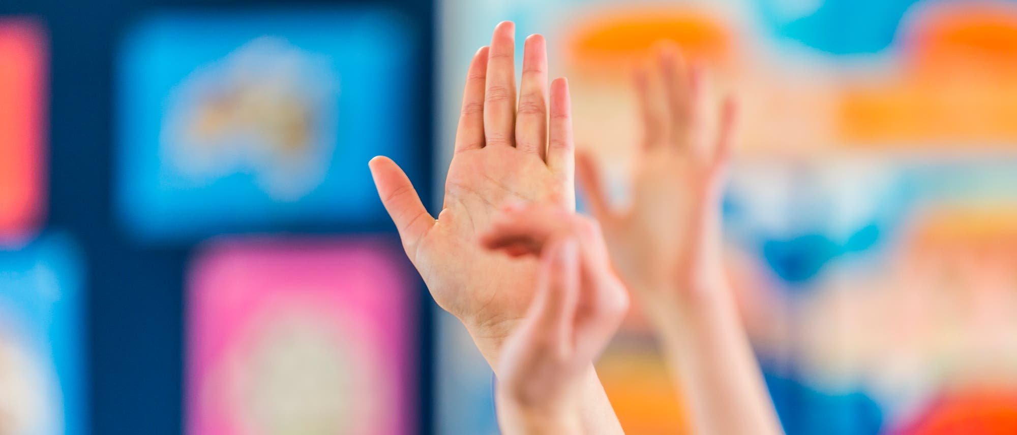 Mehrere H&auml;nde von Kindern sind in einem Klassenzimmer in die Luft gestreckt, als ob sie sich melden. Der Hintergrund ist unscharf und zeigt bunte Plakate oder Zeichnungen an der Wand.