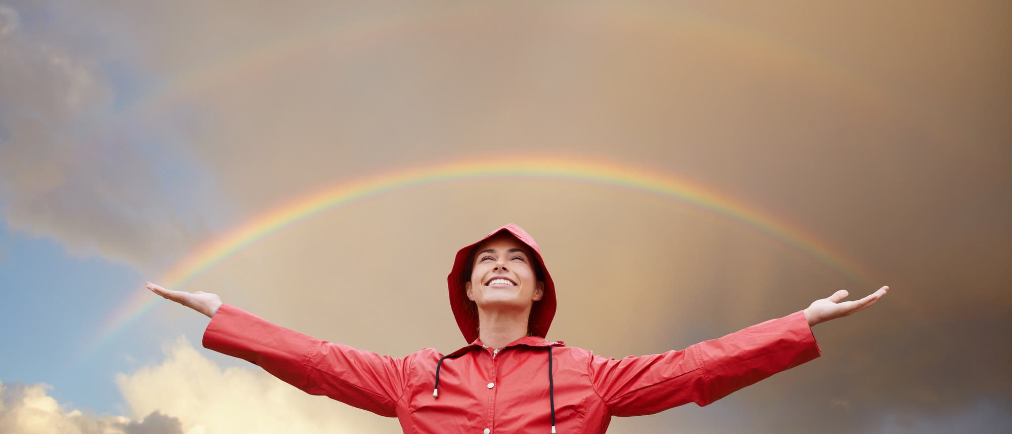 Eine Person in einem roten Regenmantel steht mit ausgebreiteten Armen vor einem doppelten Regenbogen am Himmel. Die Person l&auml;chelt und blickt nach oben. Im Hintergrund sind dramatische Wolken und ein Berg zu sehen. Die Szene vermittelt ein Gef&uuml;hl von Freude und Naturverbundenheit.