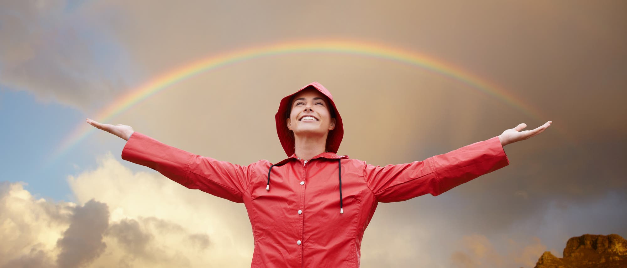 Eine Person in einem roten Regenmantel steht mit ausgebreiteten Armen vor einem Regenbogen am Himmel. Die Person lächelt und der Himmel ist teils bewölkt. Die Szene vermittelt ein Gefühl von Freude und Freiheit.