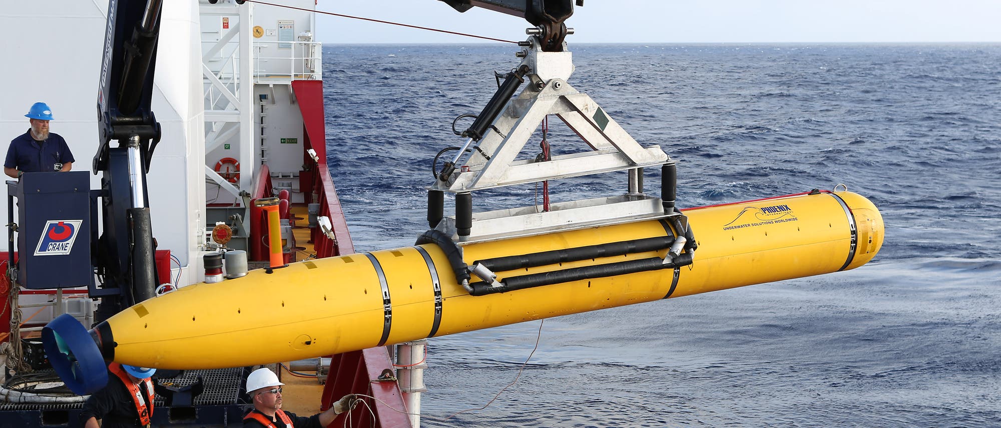 Ein gelbes Unterwasserfahrzeug wird mit einem Kran von einem Forschungsschiff ins Meer gelassen. Drei Personen in Schutzkleidung und Helmen überwachen den Vorgang. Der Ozean erstreckt sich im Hintergrund bis zum Horizont.