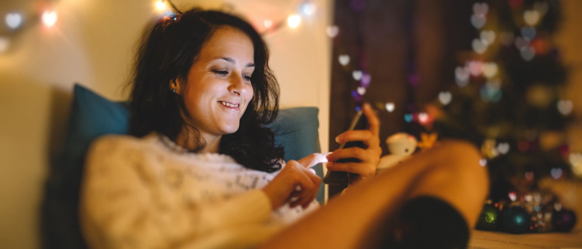 Eine Person sitzt entspannt auf einem Sofa und schaut l&auml;chelnd auf ihr Smartphone. Im Hintergrund sind bunte Lichterketten zu sehen, die eine gem&uuml;tliche Atmosph&auml;re schaffen. Die Person tr&auml;gt einen hellen Pullover und wirkt zufrieden.