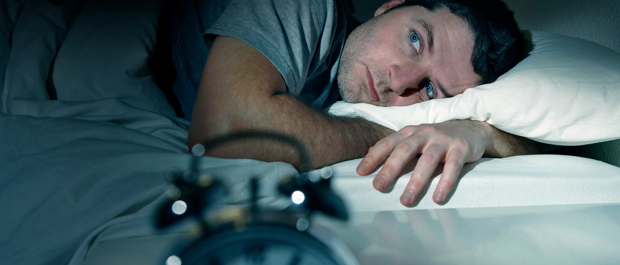 Eine Person liegt wach im Bett und schaut nachdenklich zur Seite. Im Vordergrund ist ein unscharfer Wecker zu sehen, der auf einem Nachttisch steht. Die Szene vermittelt den Eindruck von Schlaflosigkeit oder n&auml;chtlichem Gr&uuml;beln.