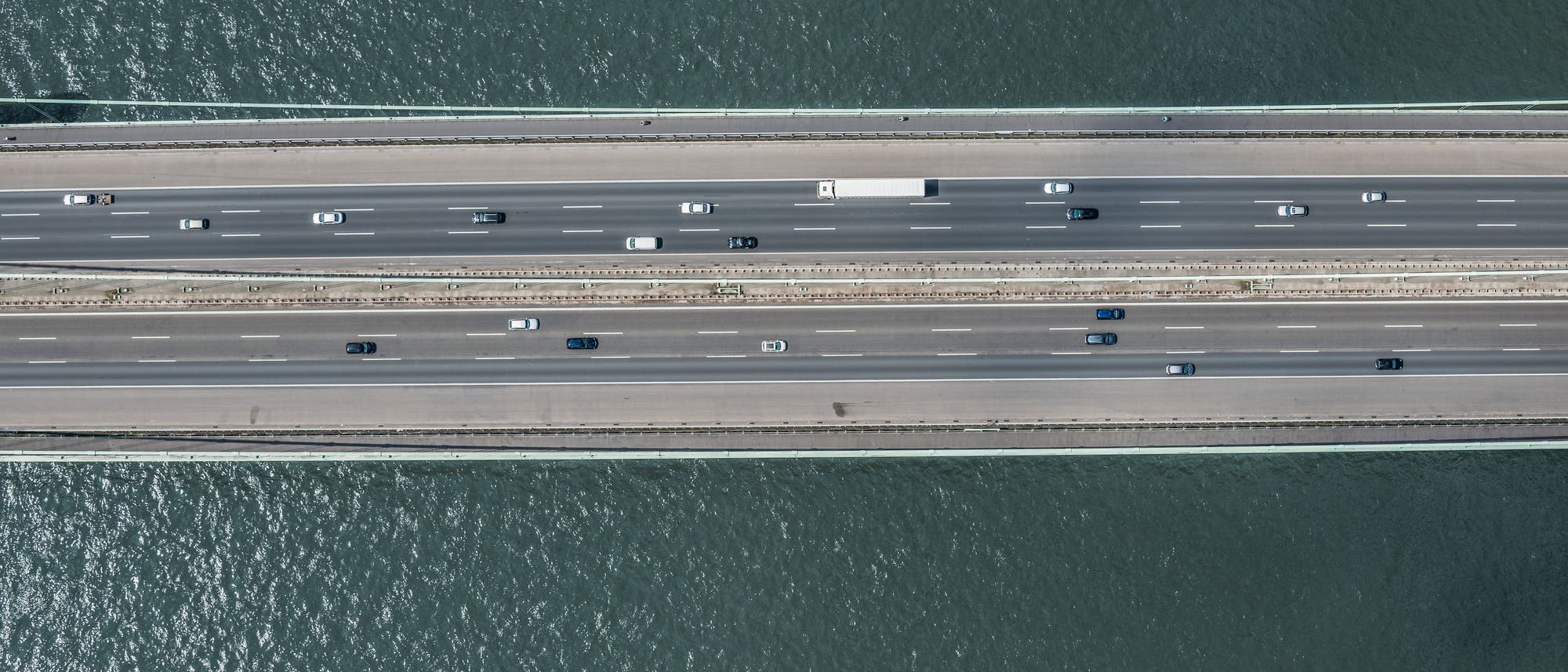 Luftaufnahme direkt von oben einer mehrspurigen Autobahnbrücke über einen Fluss. Mehrere Fahrzeuge, darunter Autos und ein Lastwagen, fahren in beide Richtungen. Die Brücke verläuft horizontal durch das Bild, während das Wasser des Flusses darunter sichtbar ist.
