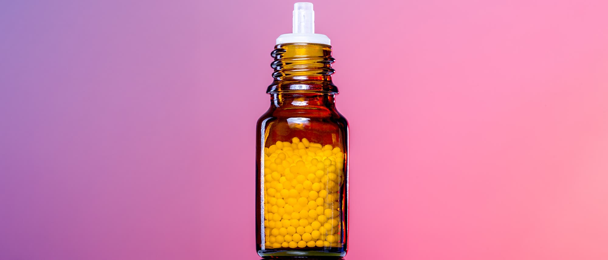 Eine braune Glasflasche mit einem weißen Verschluss, gefüllt mit homöopathischen Kügelchen. Der Hintergrund ist in einem sanften Rosa gehalten.