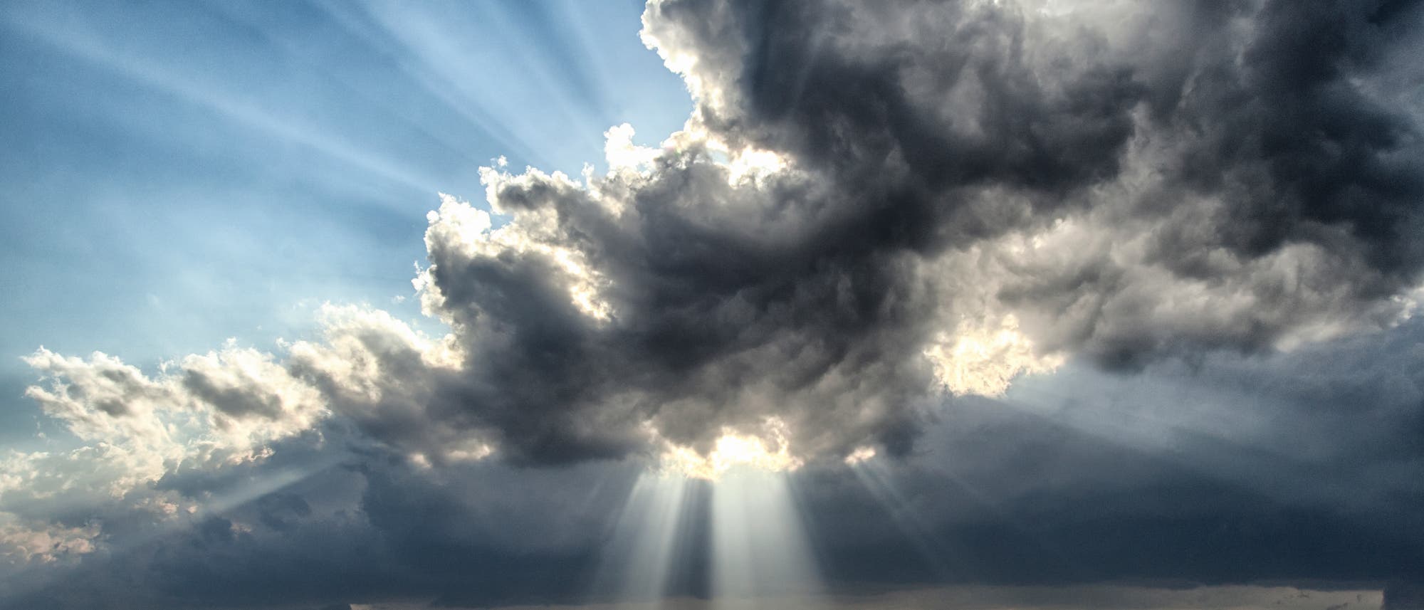 Eine weite Landschaft unter einem dramatischen Himmel, durch den Sonnenstrahlen durch dunkle Wolken brechen. Die Sonnenstrahlen erzeugen ein beeindruckendes Lichtspiel, das die darunter liegende grüne Ebene beleuchtet. Der Horizont ist flach und erstreckt sich in die Ferne, während der Himmel den Großteil des Bildes einnimmt. Eine weite Landschaft unter einem dramatischen Himmel, durch den Sonnenstrahlen durch dunkle Wolken brechen. Die Sonnenstrahlen erzeugen ein beeindruckendes Lichtspiel, das die darunter liegende grüne Ebene beleuchtet. Der Horizont ist flach und erstreckt sich in die Ferne, während der Himmel den Großteil des Bildes einnimmt.