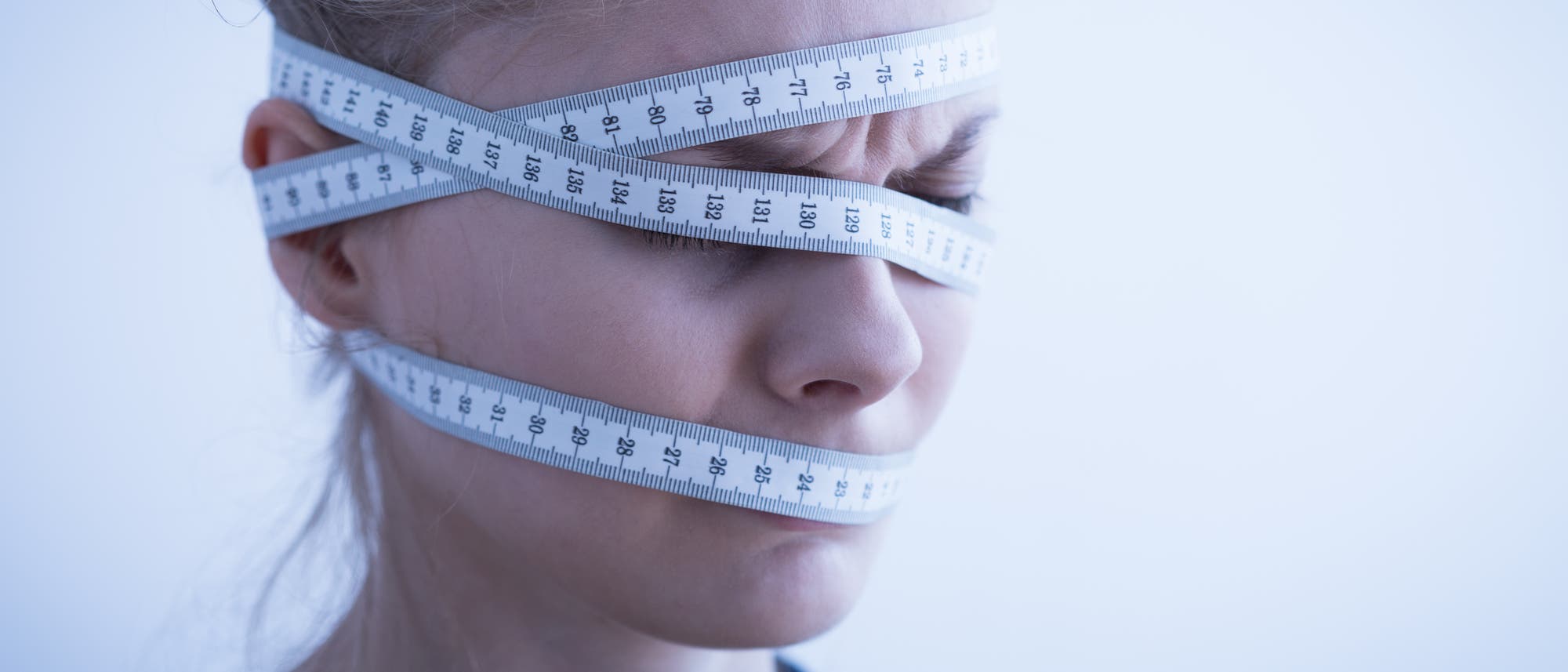 Eine Person mit geschlossenen Augen, umwickelt von einem Maßband, das über Stirn, Augen und Mund verläuft. Das Maßband zeigt Zentimeterangaben. Die Szene vermittelt ein Gefühl von Einschränkung oder Druck. Der Hintergrund ist neutral und hell. Eine Person mit geschlossenen Augen, umwickelt von einem Maßband, das über Stirn, Augen und Mund verläuft. Das Maßband zeigt Zentimeterangaben. Die Szene vermittelt ein Gefühl von Einschränkung oder Druck. Der Hintergrund ist neutral und hell.
