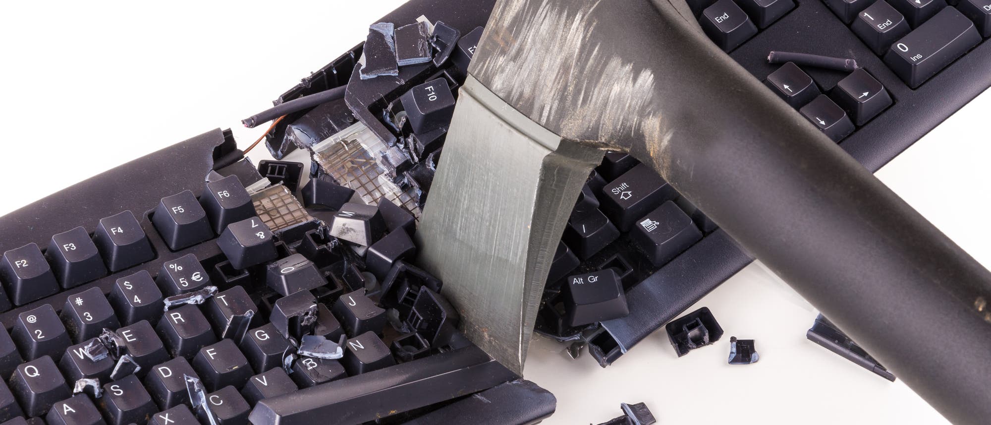 Eine schwarze Computertastatur, die mit einer Axt in der Mitte zerschlagen wurde. Tasten und Teile der Tastatur sind zerbrochen und verstreut. Die Axt ist in einem Winkel in die Tastatur eingedrungen, was auf eine gewaltsame Handlung hindeutet. Die Szene ist auf einem weißen Hintergrund dargestellt. Eine schwarze Computertastatur, die mit einer Axt in der Mitte zerschlagen wurde. Tasten und Teile der Tastatur sind zerbrochen und verstreut. Die Axt ist in einem Winkel in die Tastatur eingedrungen, was auf eine gewaltsame Handlung hindeutet. Die Szene ist auf einem weißen Hintergrund dargestellt.