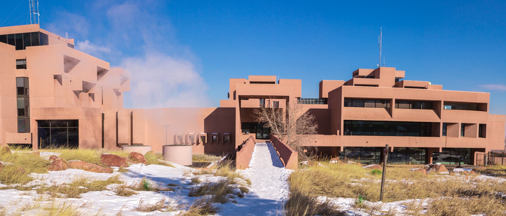 Ein modernes, mehrst&ouml;ckiges Geb&auml;ude mit einer geometrischen Architektur aus r&ouml;tlichem Beton steht in einer schneebedeckten Landschaft. Im Vordergrund sind Grasb&uuml;schel und Felsen zu sehen. Der Himmel ist klar und blau, und einige B&auml;ume flankieren das Geb&auml;ude.