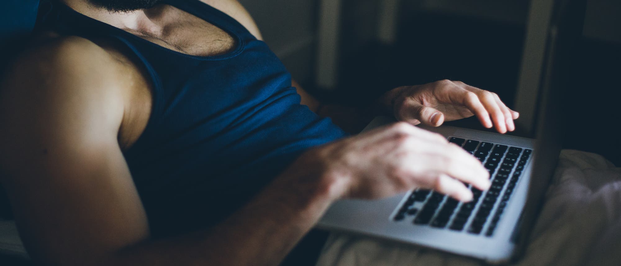 Eine Person liegt im Bett und tippt auf einem Laptop. Die Szene ist in ged&auml;mpftem Licht gehalten, was eine entspannte Atmosph&auml;re schafft. Der Fokus liegt auf den H&auml;nden, die die Tastatur bedienen. Im Hintergrund ist ein Tisch zu sehen.