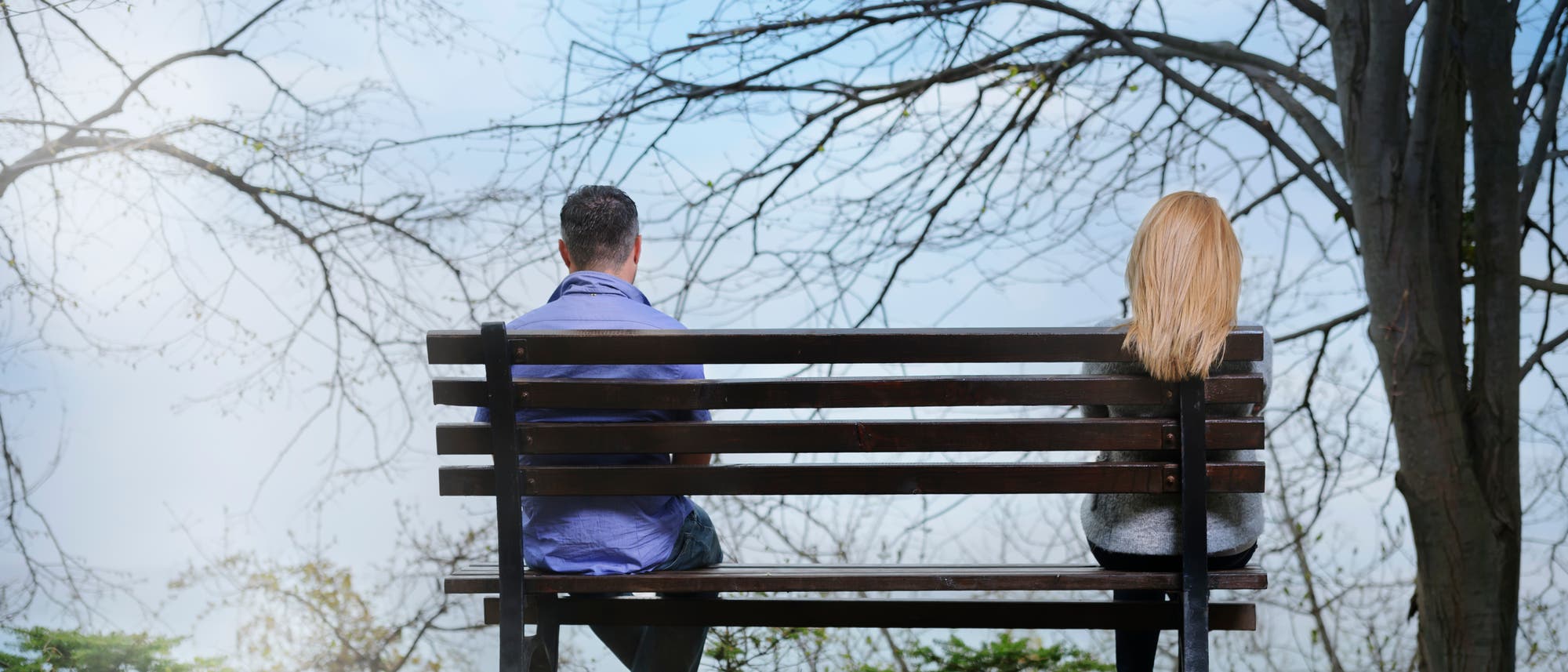 Zwei Personen sitzen weit entfernt voneinander auf einer Parkbank. Sie sind nur von hinten zu sehen: links eine Person mit kurzen dunklen Haaren, rechts eine Person mit langen blonden Haaren. Die kahlen Bäume und der wolkige Himmel verleihen der Szene eine ruhige und etwas traurige Atmosphäre. Zwei Personen sitzen weit entfernt voneinander auf einer Parkbank. Sie sind nur von hinten zu sehen: links eine Person mit kurzen dunklen Haaren, rechts eine Person mit langen blonden Haaren. Die kahlen Bäume und der wolkige Himmel verleihen der Szene eine ruhige und etwas traurige Atmosphäre.