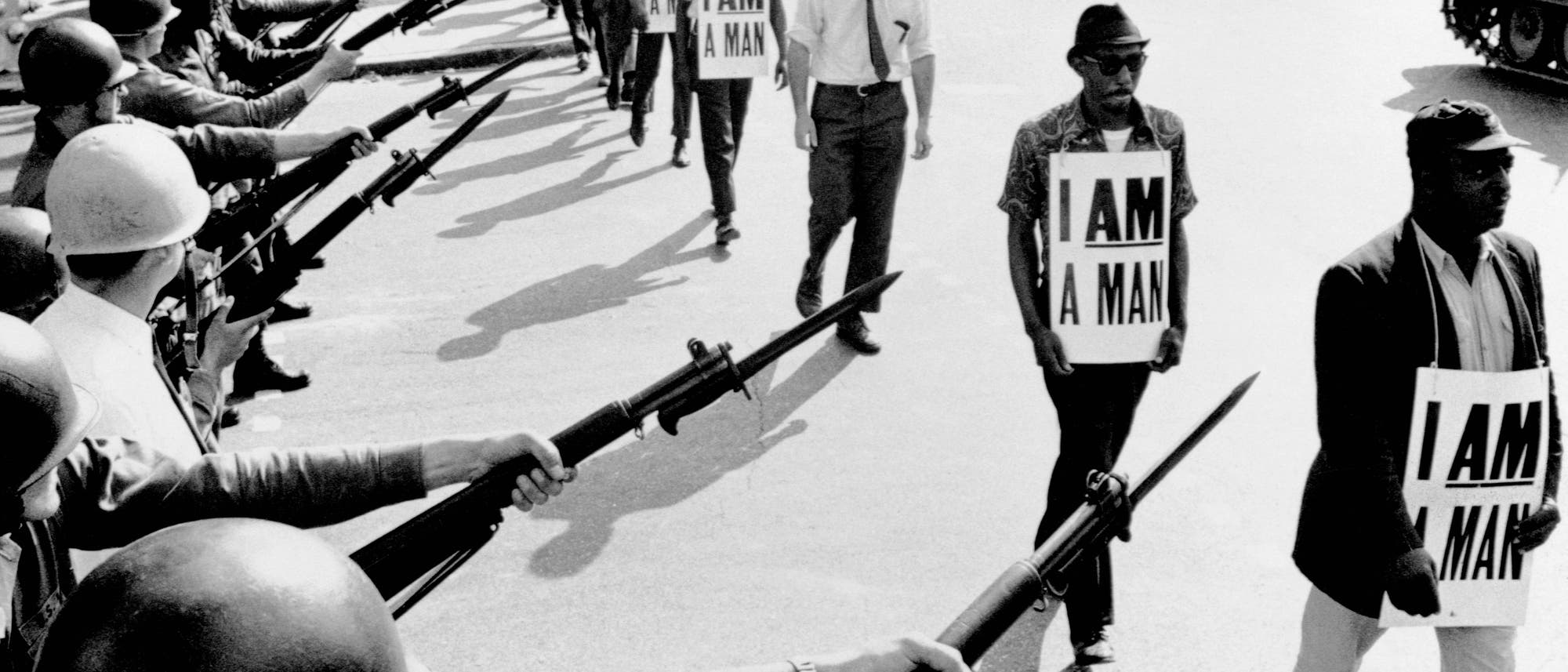 Eine Gruppe von Menschen marschiert hintereinander in einer Reihe auf einer Straße und hält Schilder mit der Aufschrift „I AM A MAN“. Ihnen gegenüber stehen Nationalgardisten mit Helmen und Gewehren. Die Szene zeigt eine angespannte Konfrontation zwischen den Demonstranten und den Gardisten. Die Demonstranten wirken entschlossen, während die Soldaten eine Barriere bilden. Das Bild vermittelt eine starke Botschaft von Protest und Widerstand. Eine Gruppe von Menschen marschiert hintereinander in einer Reihe auf einer Straße und hält Schilder mit der Aufschrift „I AM A MAN“. Ihnen gegenüber stehen Nationalgardisten mit Helmen und Gewehren. Die Szene zeigt eine angespannte Konfrontation zwischen den Demonstranten und den Gardisten. Die Demonstranten wirken entschlossen, während die Soldaten eine Barriere bilden. Das Bild vermittelt eine starke Botschaft von Protest und Widerstand.