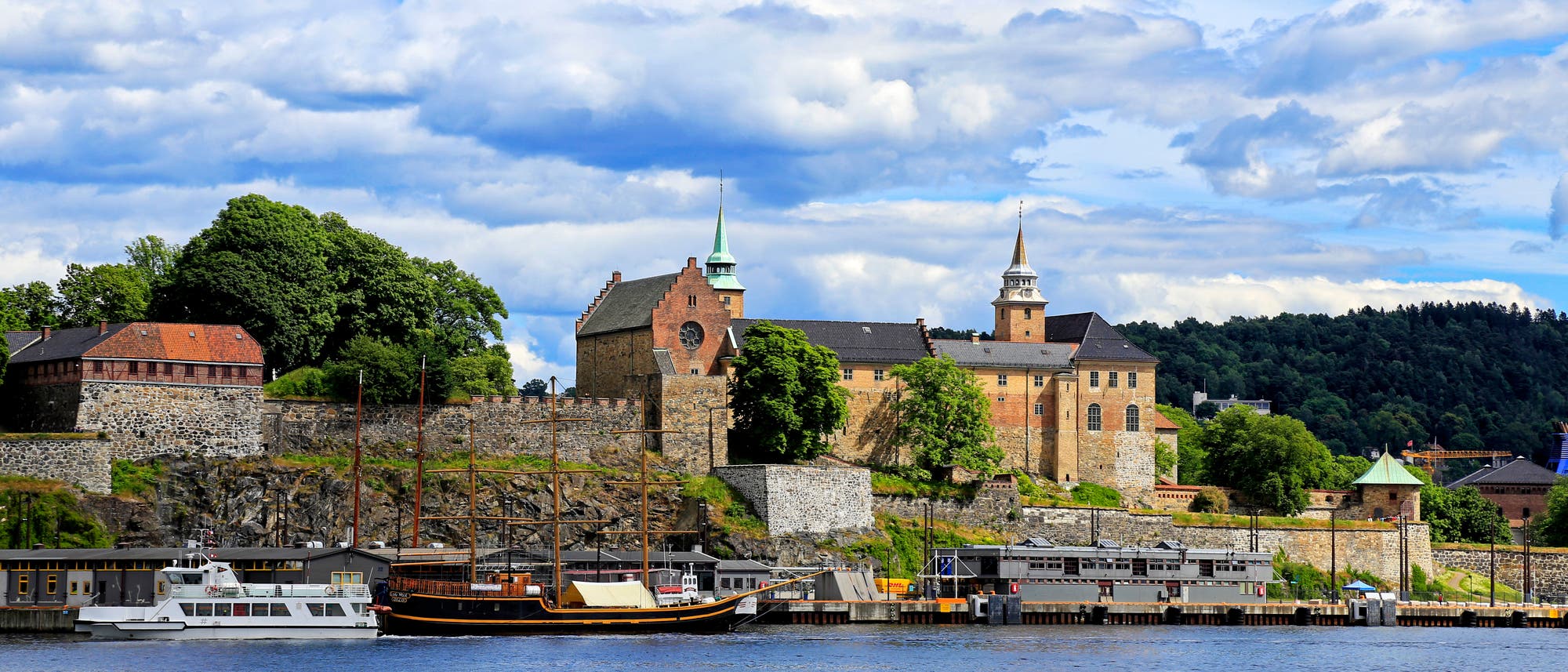Eine historische Festung mit zwei markanten T&uuml;rmen und alten Steinmauern, umgeben von &uuml;ppigem Gr&uuml;n, liegt am Ufer eines ruhigen Gew&auml;ssers. Im Vordergrund sind mehrere Boote am Kai vert&auml;ut. Der Himmel ist teils bew&ouml;lkt, was der Szene eine malerische Atmosph&auml;re verleiht.