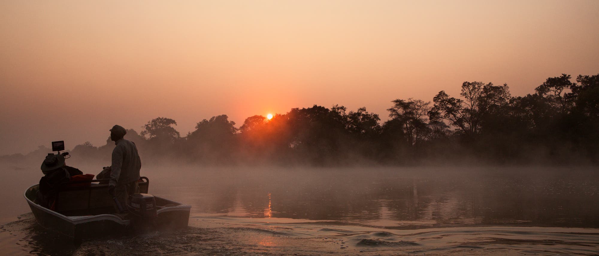 Ein Boot mit zwei Personen f&auml;hrt auf einem nebligen Fluss bei Sonnenaufgang. Die Sonne geht hinter einer Baumreihe am Horizont auf und taucht die Szene in ein warmes, orangefarbenes Licht. Die Wasseroberfl&auml;che spiegelt das Licht wider, w&auml;hrend das Boot eine leichte Welle hinterl&auml;sst.