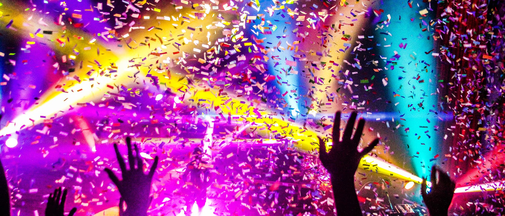 Eine lebhafte Konzertszene mit buntem Konfetti, das durch die Luft wirbelt. Mehrfarbige Lichter strahlen in den Hintergrund, w&auml;hrend H&auml;nde jubelnd in die Luft gestreckt werden. Die Atmosph&auml;re ist festlich und energiegeladen.