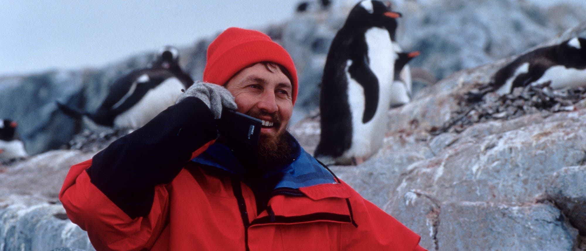 Eine Person in roter Winterkleidung und roter Mütze telefoniert lächelnd mit einem Mobiltelefon. Im Hintergrund sind mehrere Pinguine auf felsigem Gelände zu sehen. Die Szene vermittelt eine Mischung aus Abenteuer und Naturerlebnis in einer kalten, abgelegenen Umgebung. Eine Person in roter Winterkleidung und roter Mütze telefoniert lächelnd mit einem Mobiltelefon. Im Hintergrund sind mehrere Pinguine auf felsigem Gelände zu sehen. Die Szene vermittelt eine Mischung aus Abenteuer und Naturerlebnis in einer kalten, abgelegenen Umgebung.