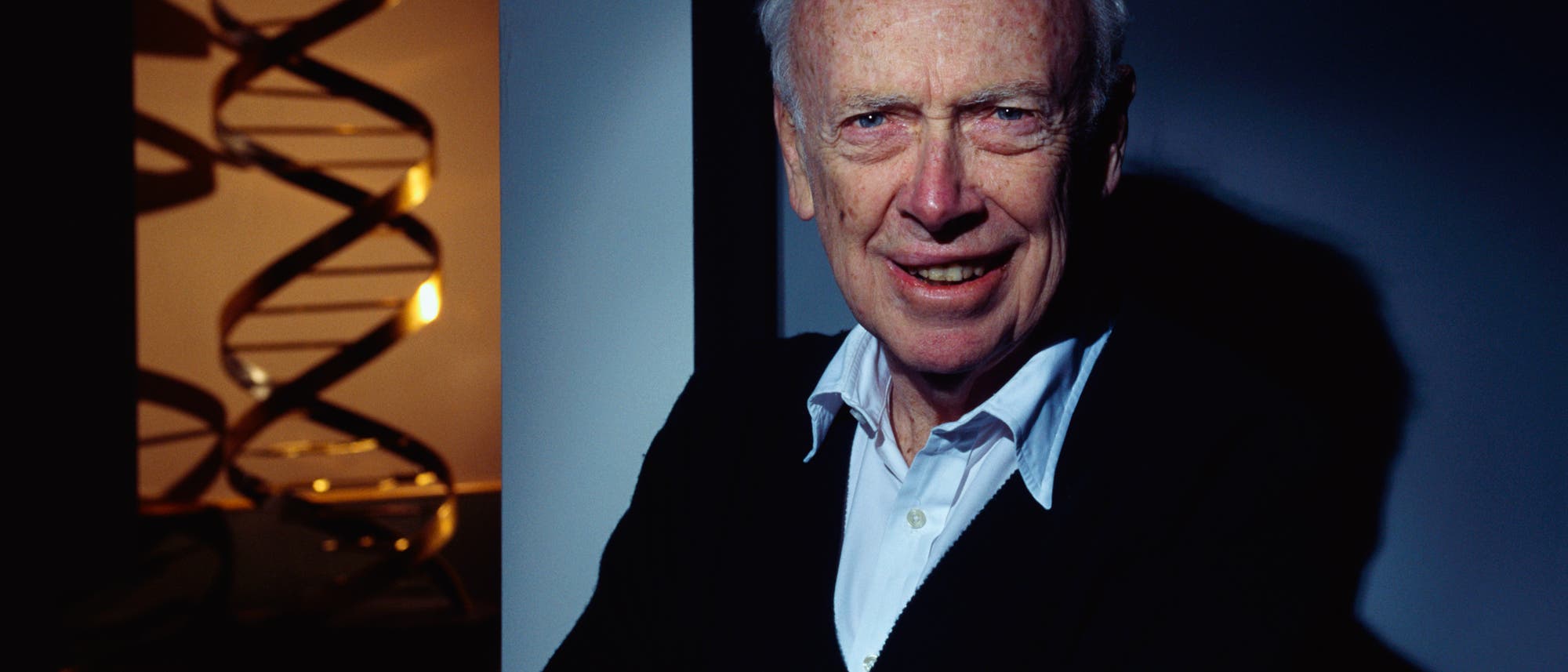 James Watson in einem weißen Hemd und dunklem Pullover steht lächelnd vor einer dunklen Kulisse. Links im Bild ist eine große, stilisierte Darstellung einer DNA-Doppelhelix zu sehen, die im Licht schimmert. Die Szene vermittelt eine wissenschaftliche Atmosphäre. James Watson in einem weißen Hemd und dunklem Pullover steht lächelnd vor einer dunklen Kulisse. Links im Bild ist eine große, stilisierte Darstellung einer DNA-Doppelhelix zu sehen, die im Licht schimmert. Die Szene vermittelt eine wissenschaftliche Atmosphäre.