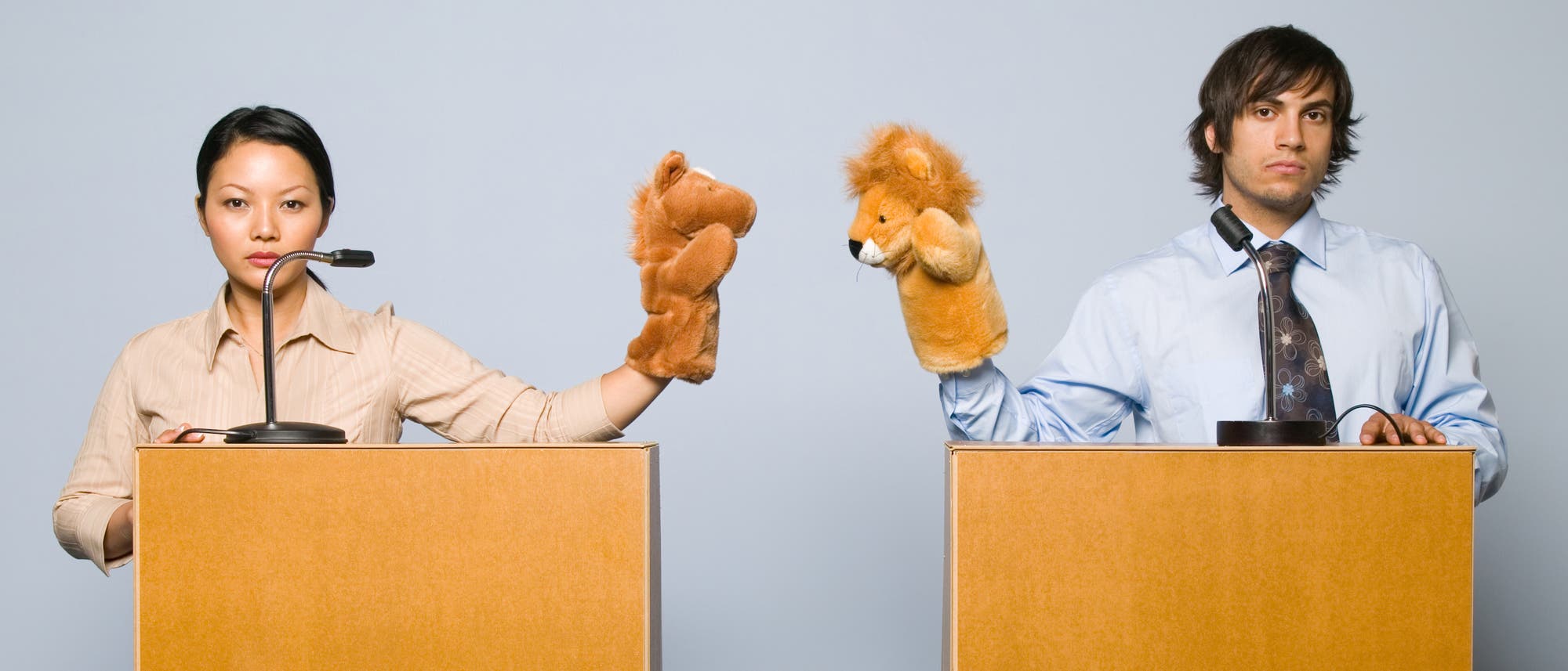 Zwei Personen stehen nebeneinander hinter Rednerpulten und halten Handpuppen. Die Frau links hält eine Pferdepuppe, der Mann rechts eine Löwenpuppe. Die Puppen sind aufeinander gerichtet, die Personen schauen sich demonstrativ nicht an, sondern ernst in die Kamera. Obwohl vor beiden ein Mikro steht, haben sie den Mund geschlossen. Der Hintergrund ist neutral grau.