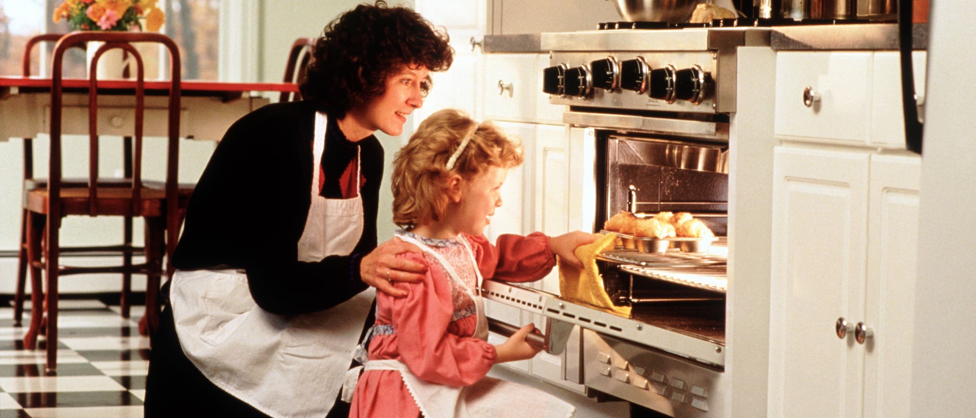 Eine Frau und ein Kind, beide in Schürzen, stehen in einer Küche vor einem geöffneten Backofen. Frisuren und Kleidung der beiden lassen darauf schließen, dass die Aufnahme bereits etwas älter ist. Die erwachsene Frau legt dem Kind sanft die Hand auf die Schulter, während es ein Backblech mit Gebäck in den Ofen schiebt. Im Hintergrund ist ein Tisch mit Blumen zu sehen. Die Szene vermittelt eine warme, häusliche Atmosphäre.