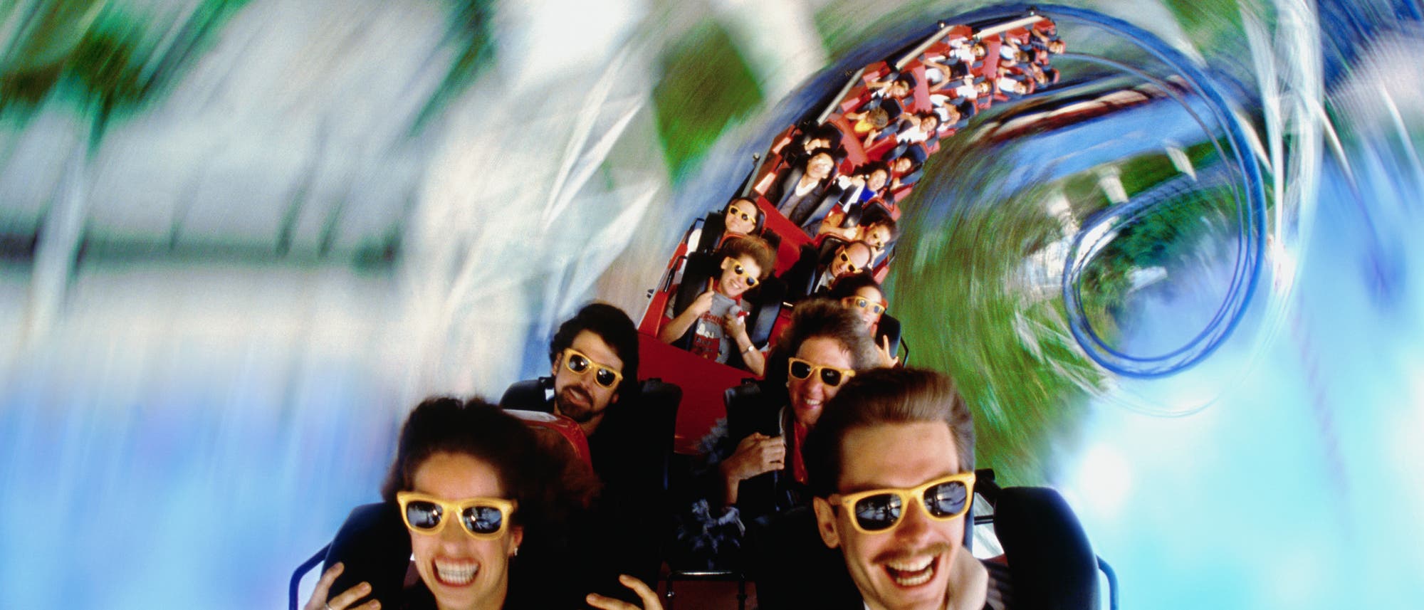 Menschen fahren in einem Achterbahnwagen durch eine Looping-Strecke. Sie tragen Sonnenbrillen und wirken begeistert und aufgeregt. Die Umgebung ist verschwommen, was die Geschwindigkeit und Dynamik der Fahrt betont. Die Szene vermittelt ein Gefühl von Abenteuer und Spaß.