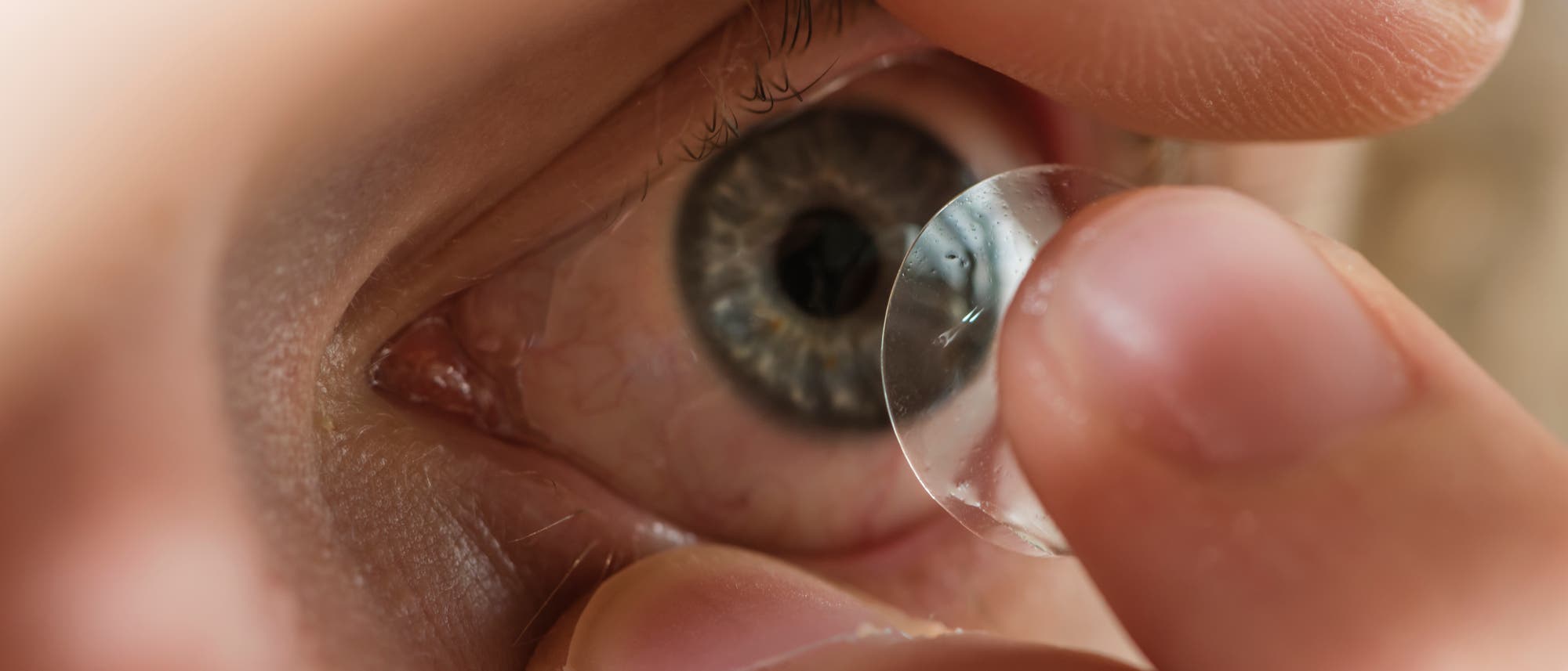 Eine Person setzt eine Kontaktlinse auf ein Auge. Die Nahaufnahme zeigt die Linse, die mit den Fingerspitzen gehalten wird, während das Auge weit geöffnet ist. Die Details der Iris sind deutlich sichtbar. Eine Person setzt eine Kontaktlinse auf ein Auge. Die Nahaufnahme zeigt die Linse, die mit den Fingerspitzen gehalten wird, während das Auge weit geöffnet ist. Die Details der Iris sind deutlich sichtbar.