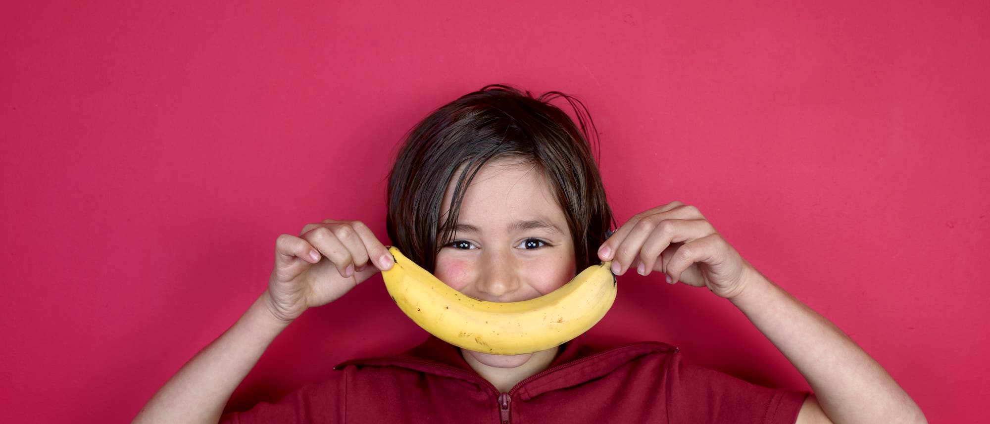 Ein Junge vor pinkfarbenem Hintergrund hält eine Banane vor seinen Mund, so dass sie sein Lächeln ergibt. 