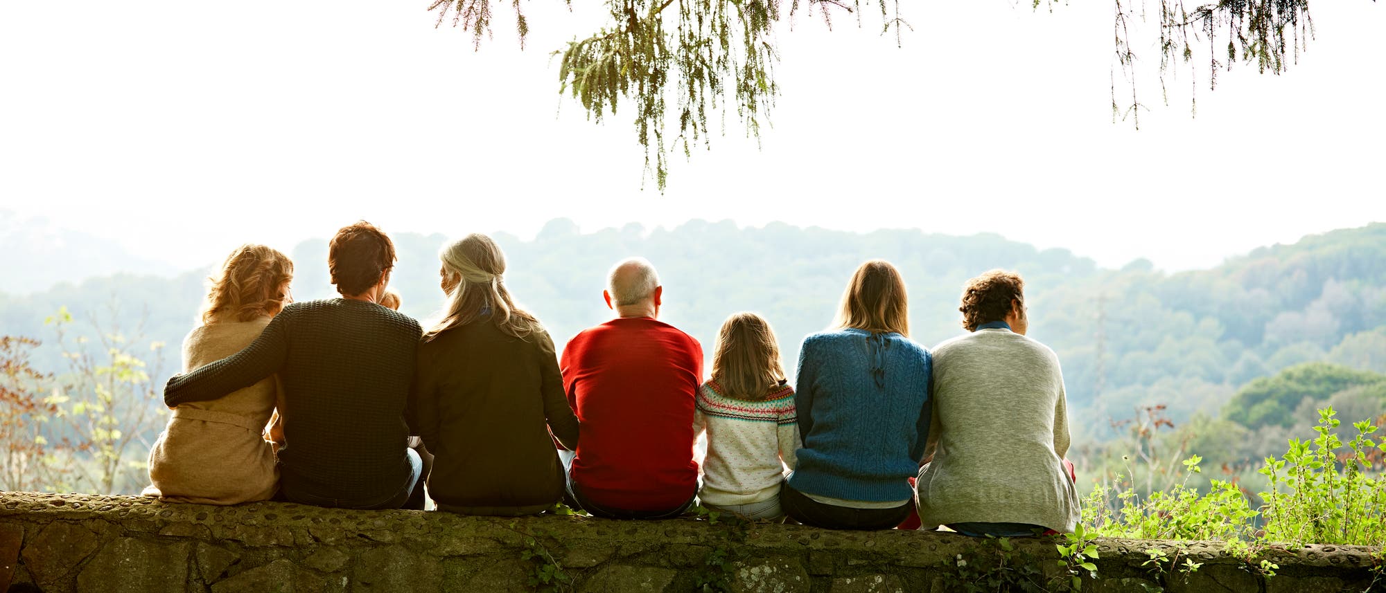 Eine Gruppe von sieben Personen unterschiedlichen Alters sitzt auf einer Steinmauer und blickt auf eine bewaldete Landschaft. Im Vordergrund ist grünes Gras zu sehen, während im Hintergrund Bäume und Hügel die Aussicht prägen. Eine Gruppe von sieben Personen unterschiedlichen Alters sitzt auf einer Steinmauer und blickt auf eine bewaldete Landschaft. Im Vordergrund ist grünes Gras zu sehen, während im Hintergrund Bäume und Hügel die Aussicht prägen.