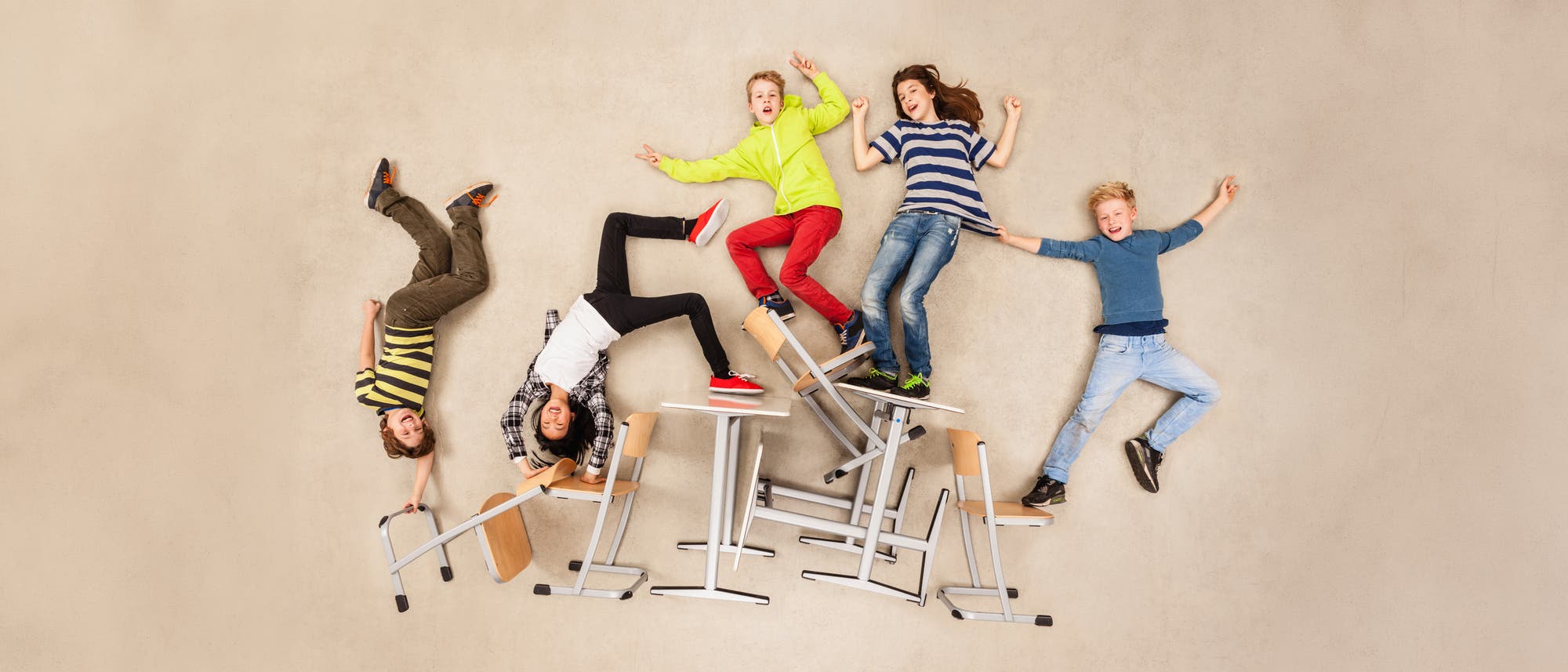 Eine Gruppe von f&uuml;nf Sch&uuml;lerinnen und Sch&uuml;lern liegt auf dem Boden und posiert fr&ouml;hlich, als ob sie an einer Wand h&auml;ngen w&uuml;rden. Sie sind von umgekippten St&uuml;hlen und Tischen umgeben, was eine spielerische und dynamische Szene erzeugt. Die Kinder tragen bunte Freizeitkleidung und wirken ausgelassen und energiegeladen. Die Szene vermittelt eine Atmosph&auml;re von Schule und Aufbruch.