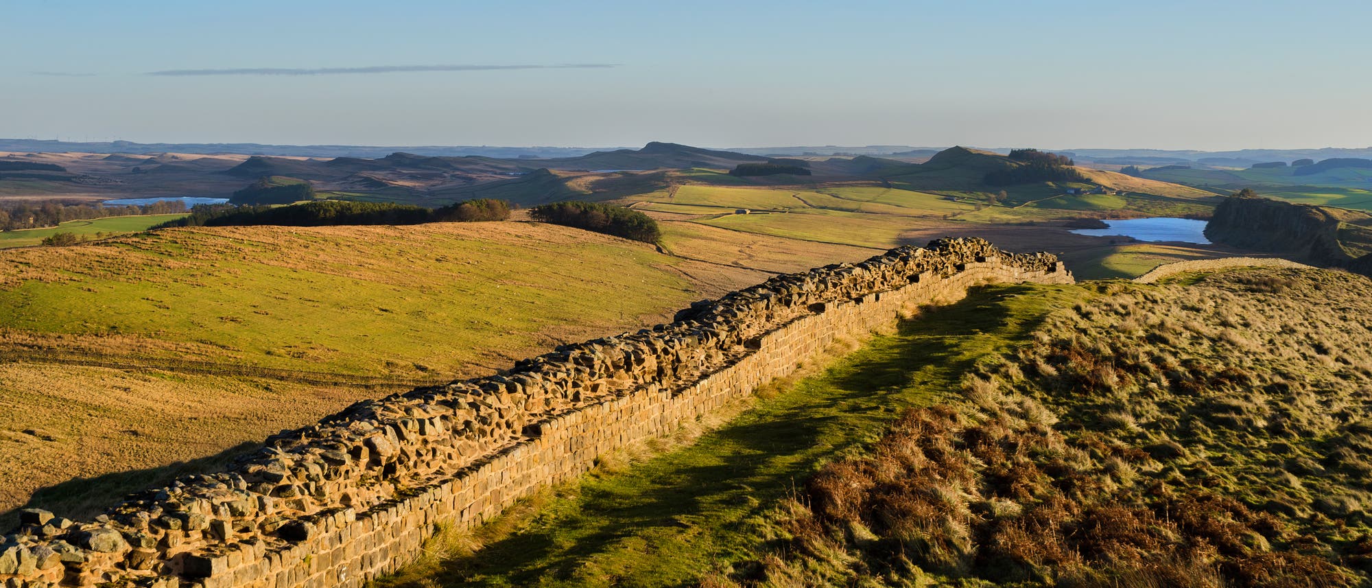 Eine weitl&auml;ufige Landschaft mit sanften H&uuml;geln und einer historischen Steinmauer, die sich durch die Szene zieht. Im Hintergrund sind ein See und bewaldete Bereiche zu sehen. Der Himmel ist klar und blau, was auf einen sonnigen Tag hindeutet.