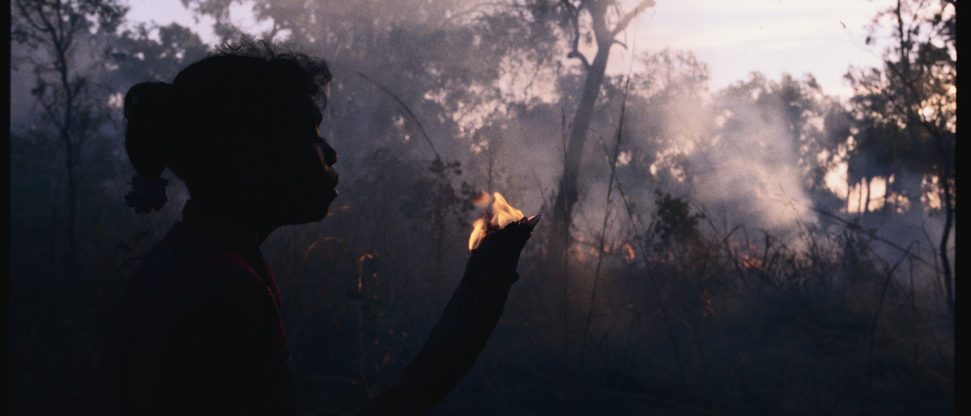 Eine Angehörige eines indigenen Volkes in Australien hält einen brennenden Ast. In Hintergrund lodern Flammen im Unterholz.