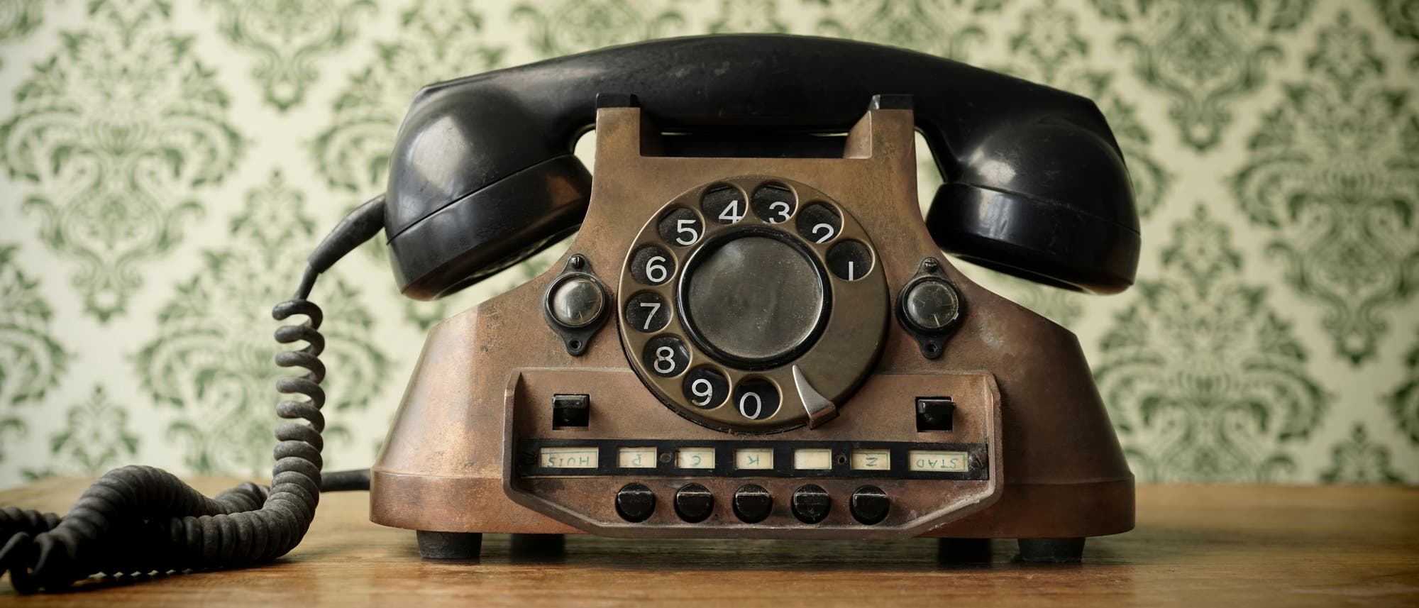 Ein altes, braunes W&auml;hlscheibentelefon mit schwarzem H&ouml;rer steht auf einem Holztisch. Die W&auml;hlscheibe zeigt die Zahlen 1 bis 0. Im Hintergrund ist eine gr&uuml;n-wei&szlig;e Tapete mit einem floralen Muster zu sehen. Das Telefon hat mehrere Tasten unterhalb der W&auml;hlscheibe.