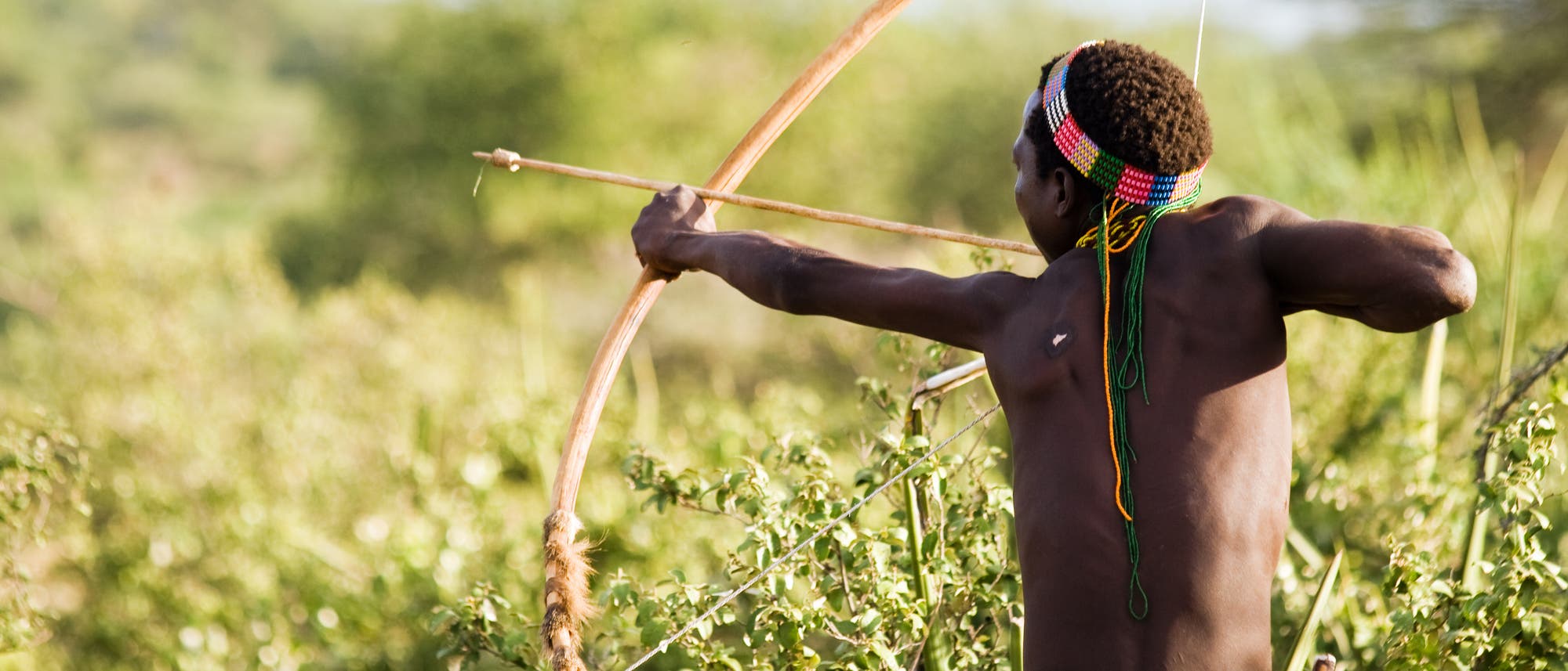 Eine Person steht in einer natürlichen Umgebung und spannt einen Bogen, bereit, einen Pfeil abzuschießen. Sie trägt ein buntes Stirnband und hat den Blick auf das Ziel gerichtet. Im Hintergrund ist dichte Vegetation zu sehen, die eine ländliche oder wilde Landschaft andeutet.