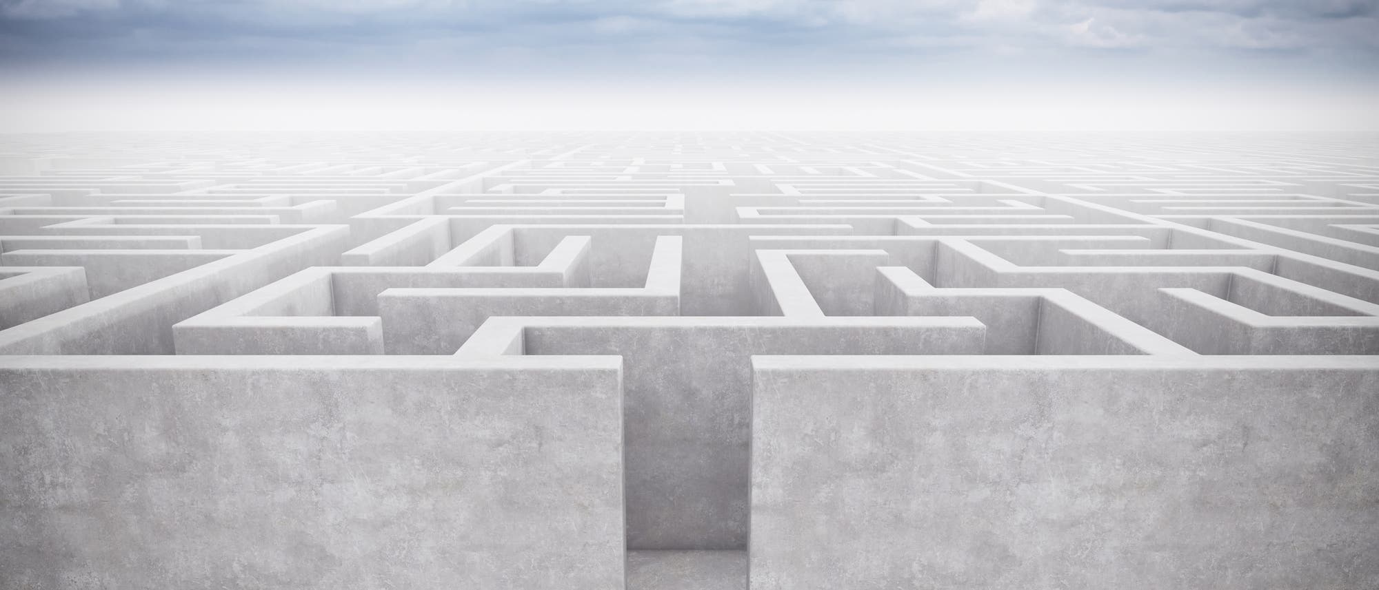 Eine Person in einem Anzug steht vor einem großen, komplexen Labyrinth aus hohen, grauen Wänden, das sich bis zum Horizont erstreckt. Der Himmel ist bewölkt, was eine nachdenkliche und herausfordernde Atmosphäre schafft. Die Person hält einen Aktenkoffer und scheint bereit, das Labyrinth zu betreten, was symbolisch für eine bevorstehende Herausforderung oder Entscheidung steht.