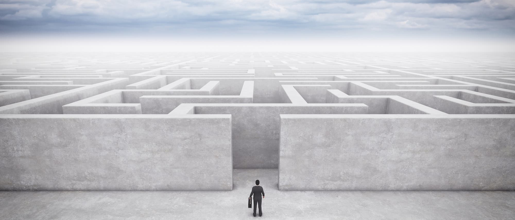 Eine Person in einem Anzug steht vor einem großen, komplexen Labyrinth aus hohen, grauen Wänden, das sich bis zum Horizont erstreckt. Der Himmel ist bewölkt, was eine nachdenkliche und herausfordernde Atmosphäre schafft. Die Person hält einen Aktenkoffer und scheint bereit, das Labyrinth zu betreten, was symbolisch für eine bevorstehende Herausforderung oder Entscheidung steht.