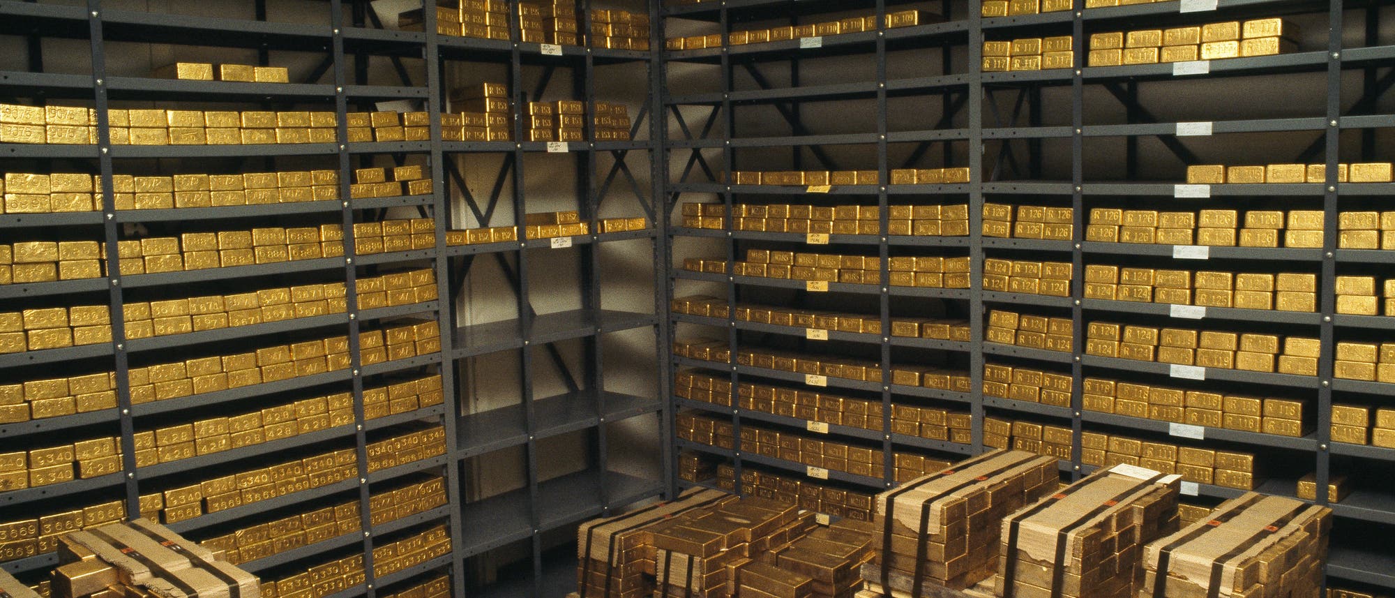 Ein Raum mit Regalen voller gestapelter Goldbarren. Die Barren sind in mehreren Reihen und Spalten angeordnet, sowohl auf den Regalen als auch auf Paletten am Boden. Die Szene vermittelt den Eindruck eines Tresorraums oder einer Lagerst&auml;tte f&uuml;r wertvolle Metalle.