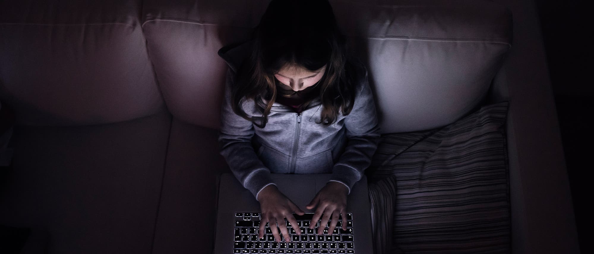 Ein Mädchen sitzt in einem dunklen Raum auf einem Sofa und tippt auf einem Laptop. Die Szene vermittelt eine düstere, einsame Atmosphäre.